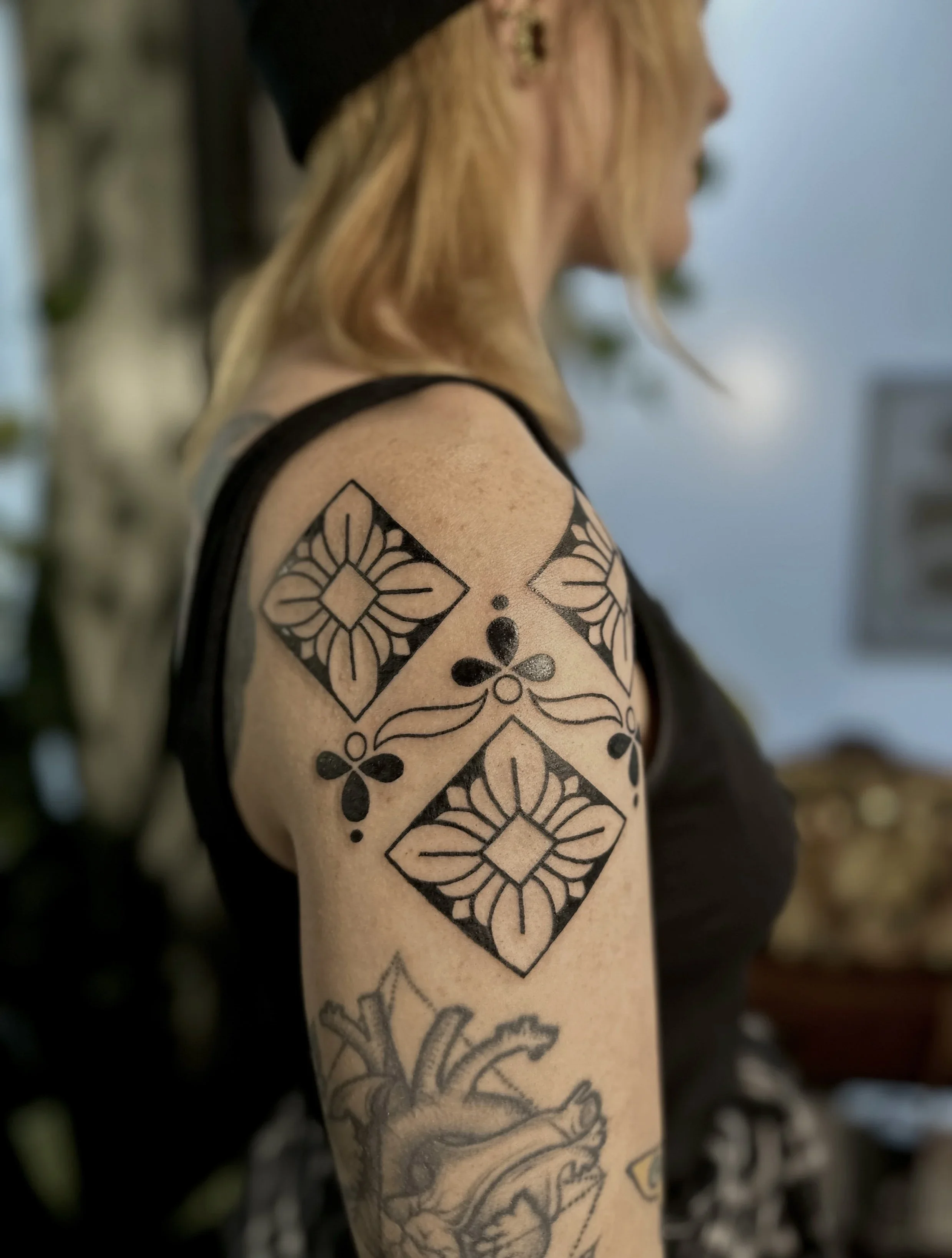 Part of a woman's upper arm with a black geometric tattoo, including diamond and floral patterns, and blonde hair with black hat visible.