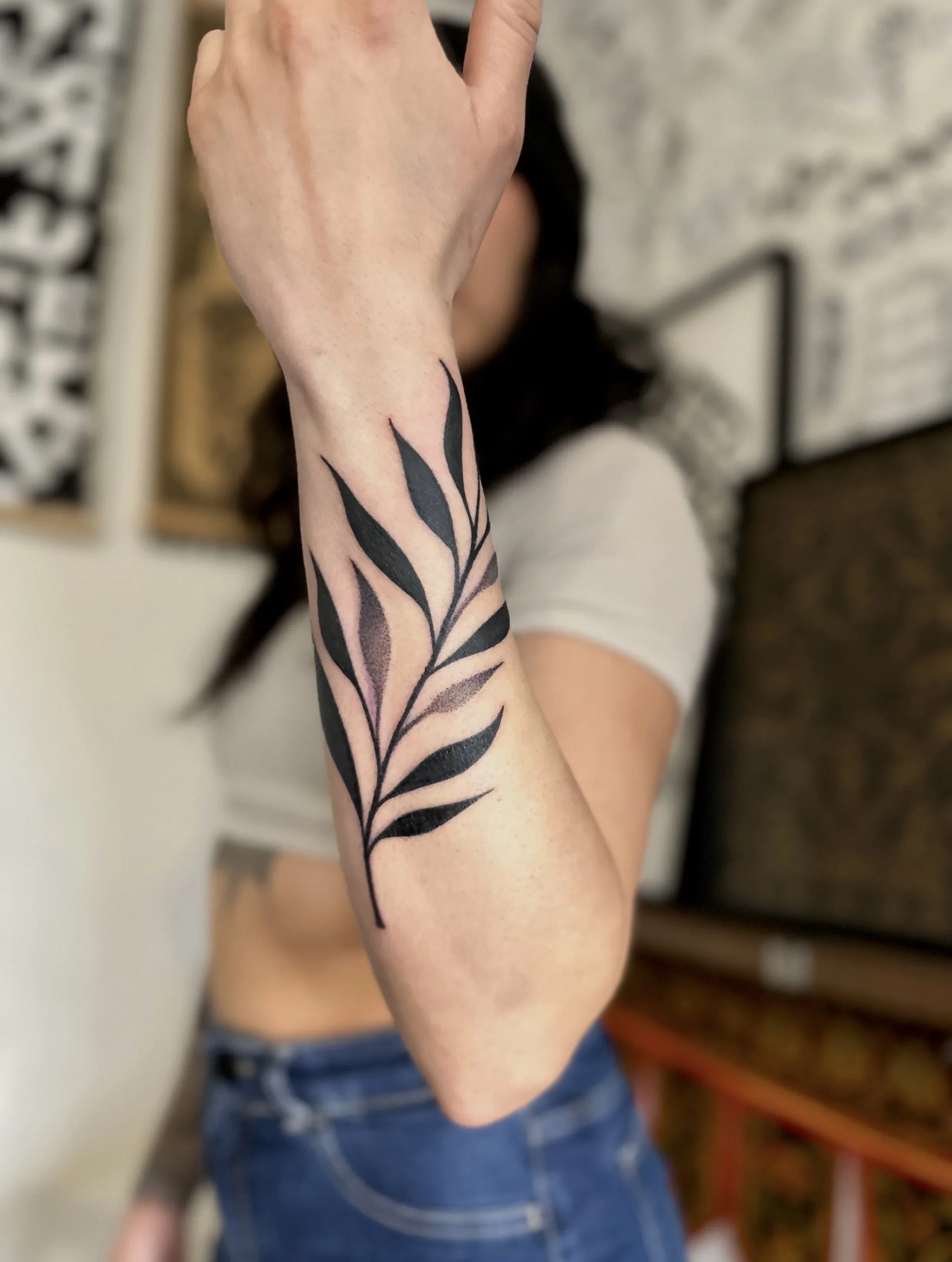Close-up of a person's arm with a black tattoo of elongated leaves on the forearm. The person is wearing a short-sleeved shirt, and the background includes a blurred person wearing blue jeans and a white top.