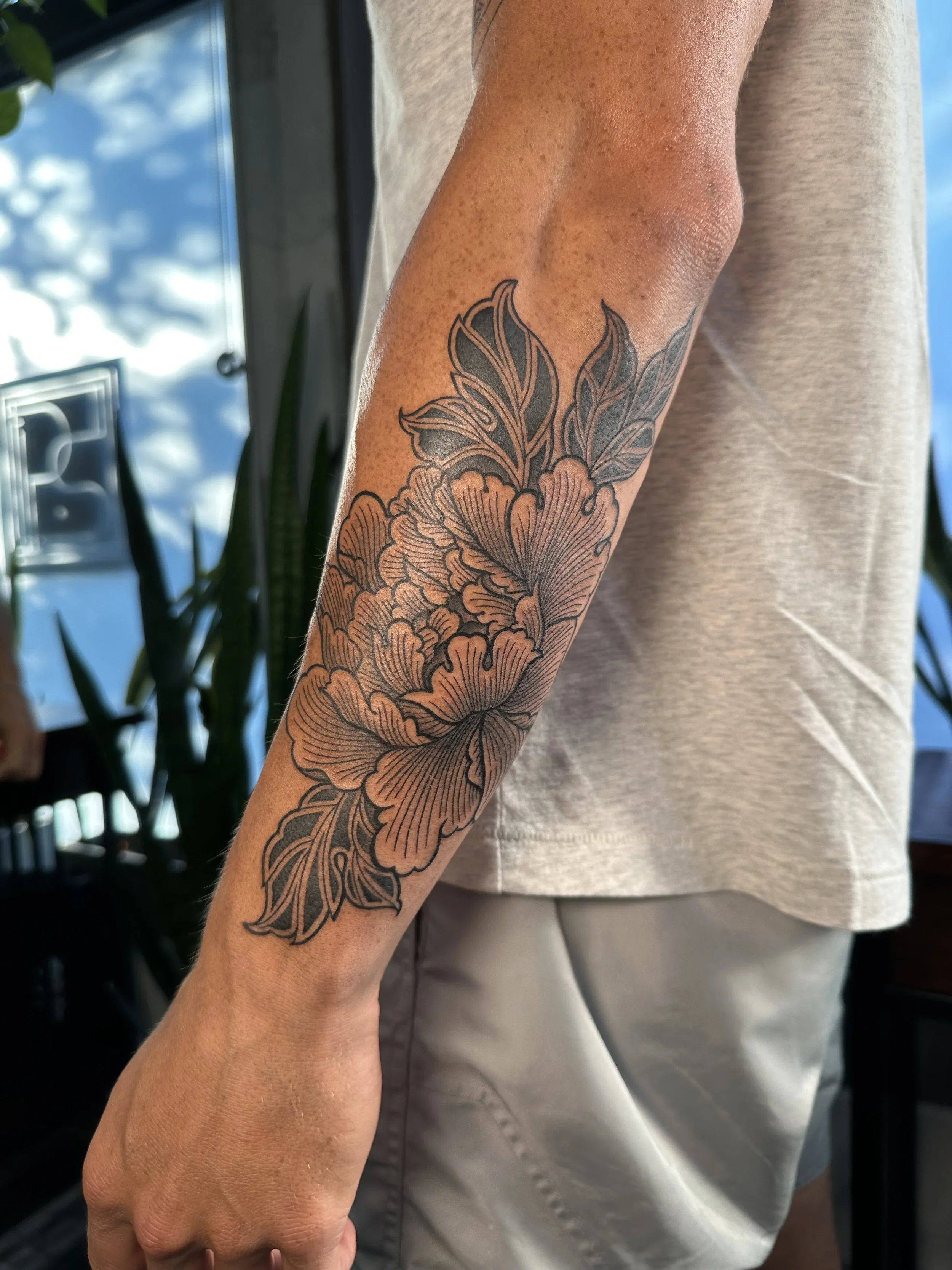 A person's arm with a black and gray floral tattoo extending from the wrist to the elbow, wearing a light gray shirt, with a background of a window, plant, and cloudy sky.