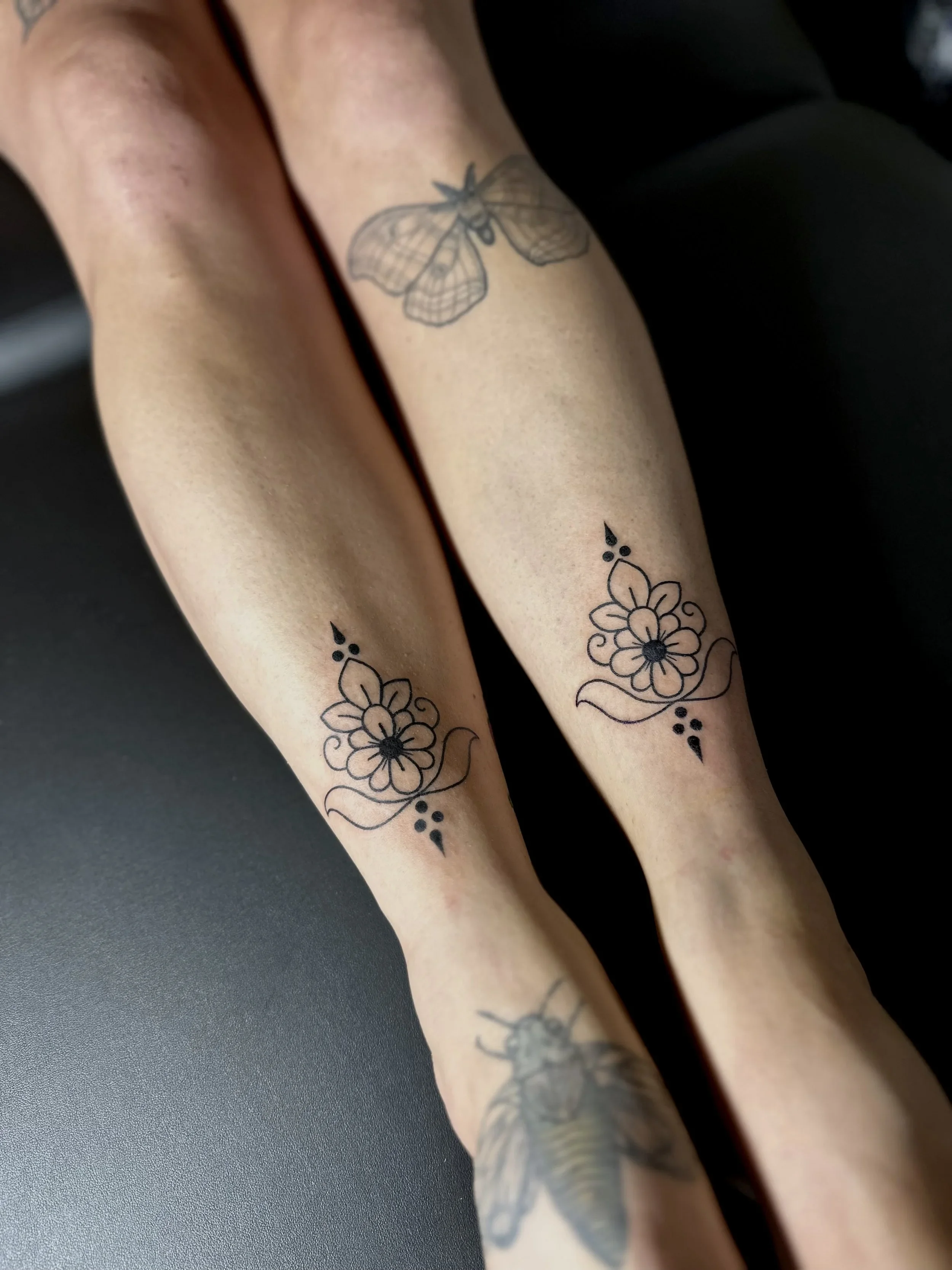 Close-up of a person's arms with black line art tattoos, including a moth and floral designs.