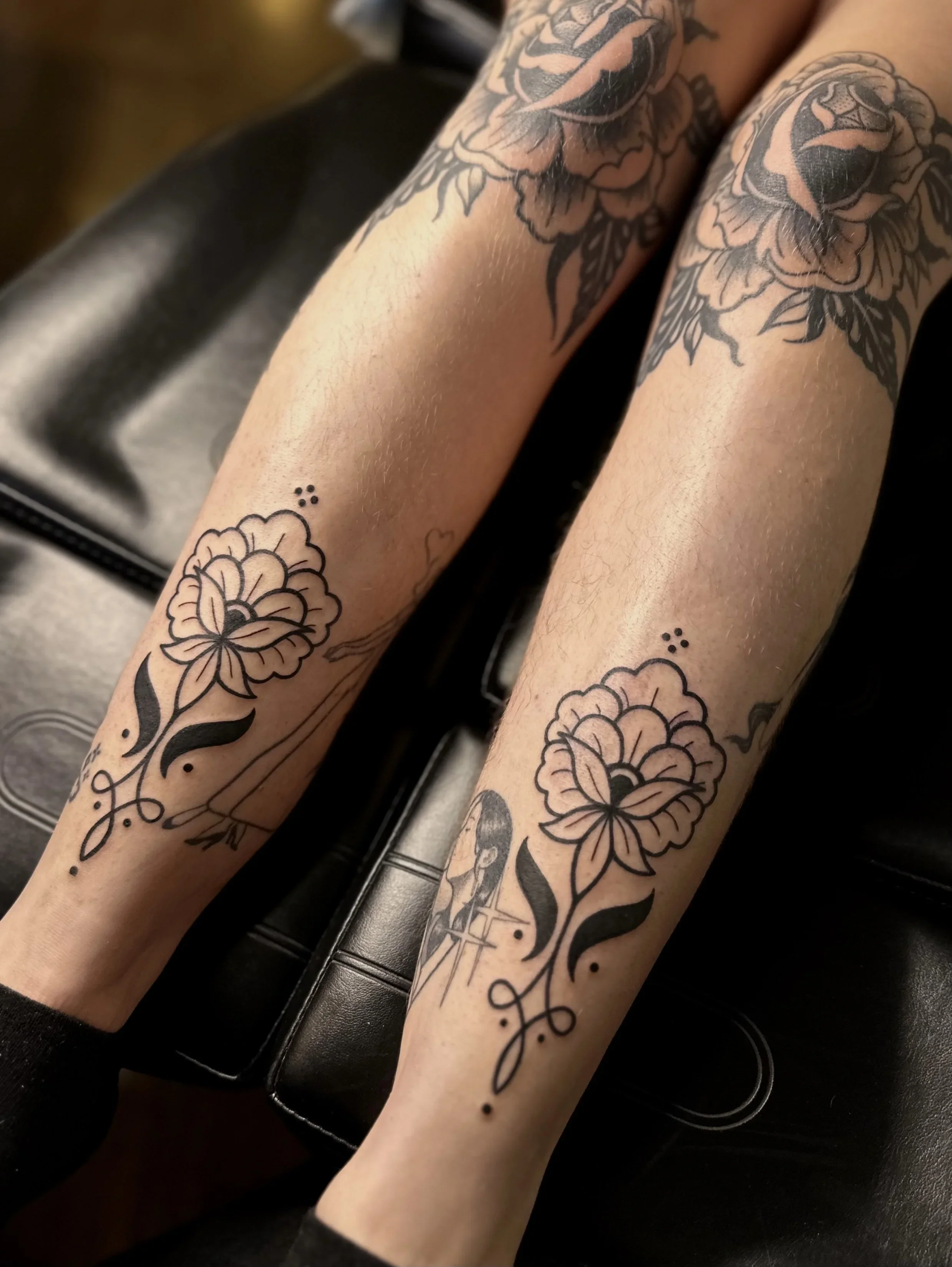 Close-up of two legs with black ink floral tattoos, resting on a black leather surface.
