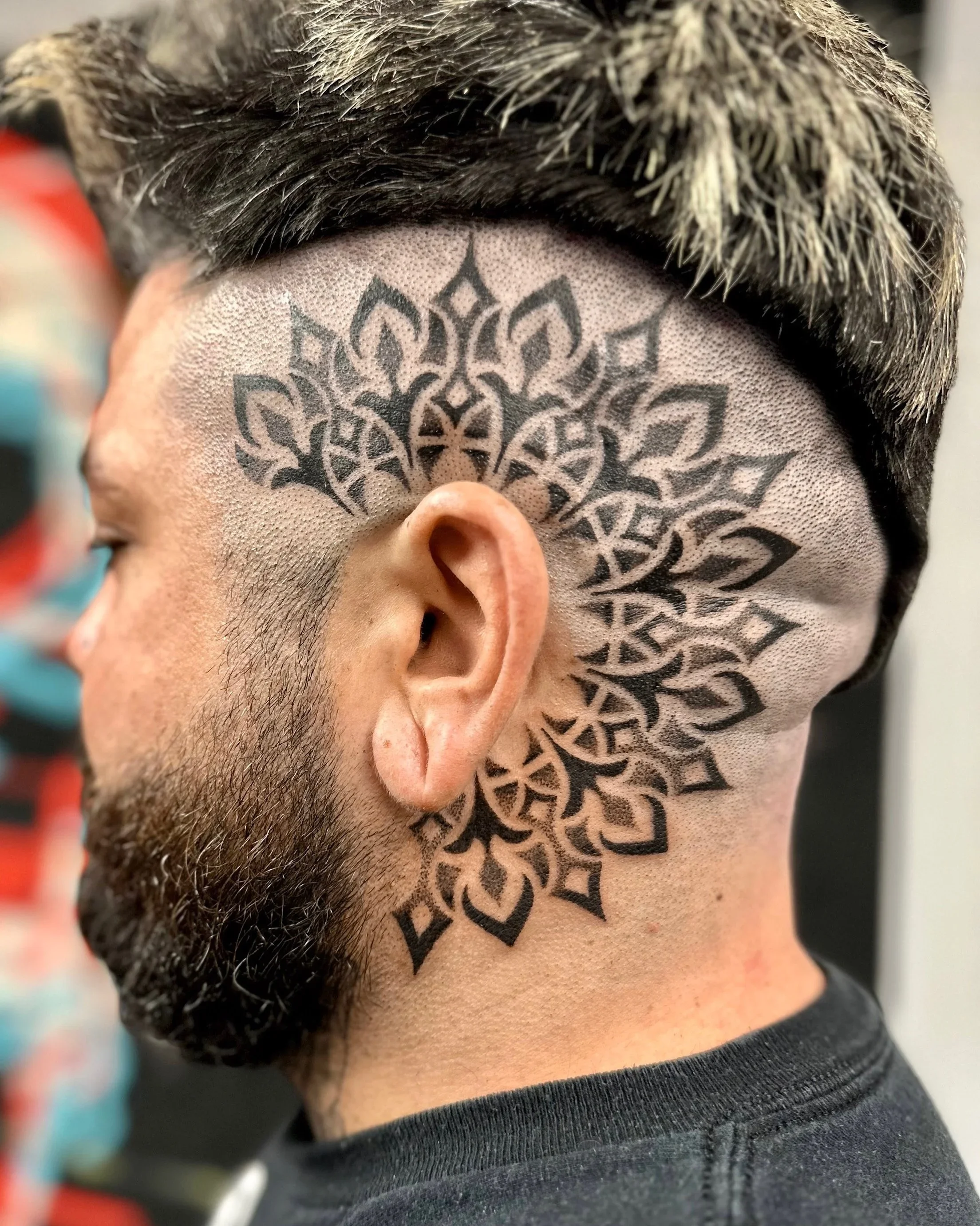 Close-up of a tattoo behind an ear depicting a black tribal mandala pattern on the scalp.