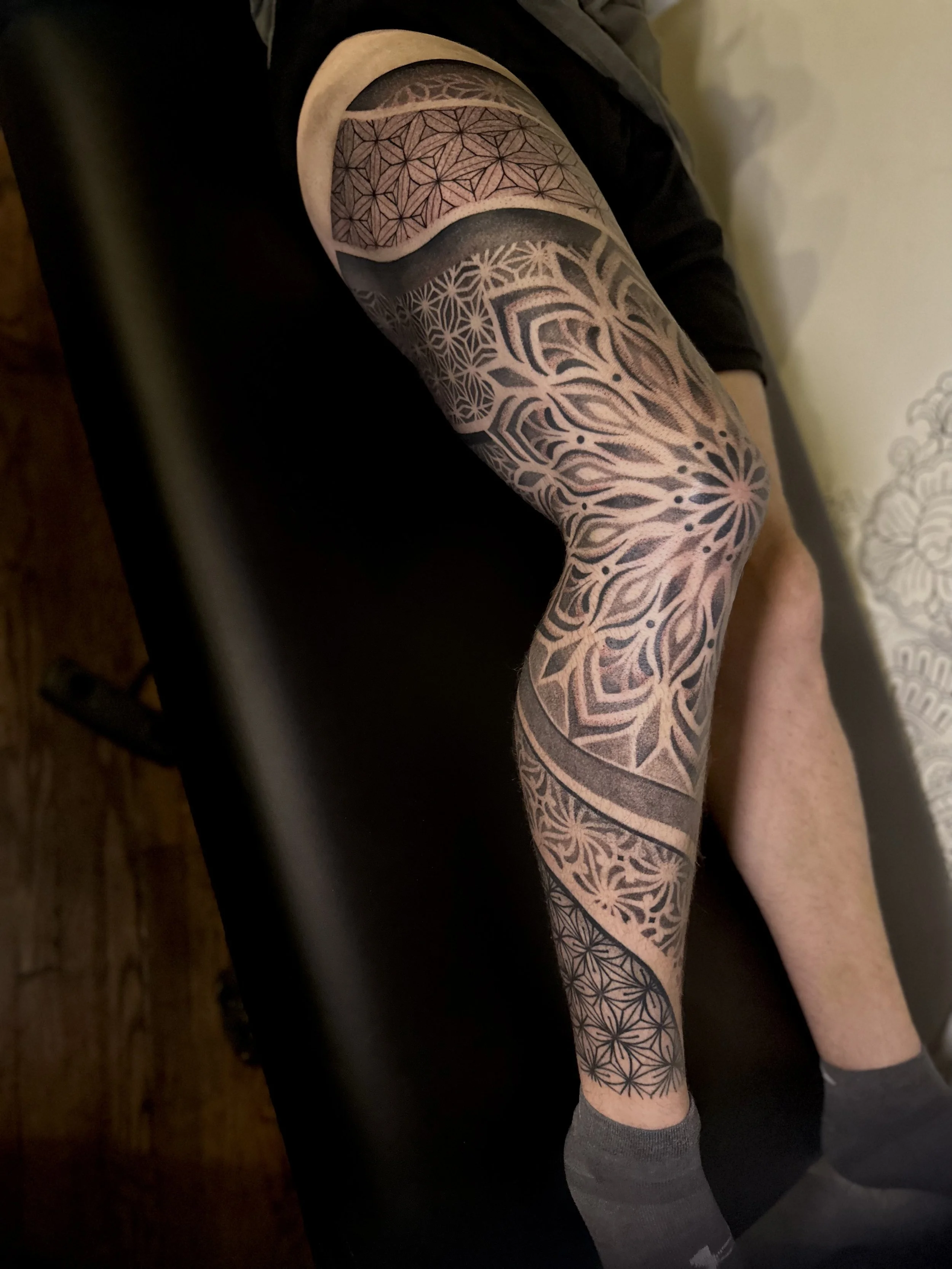 Close-up of a person's arm with a large, intricate geometric tattoo covering the upper arm and forearm, featuring various patterns and black and gray shading.