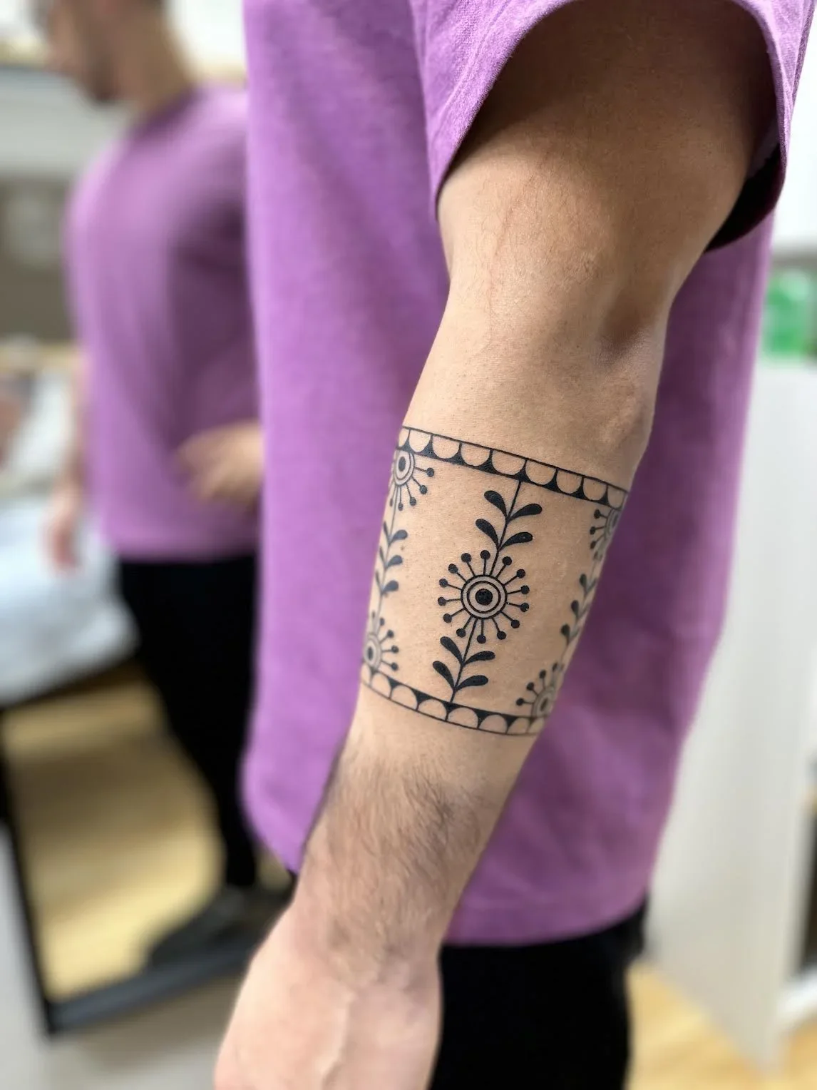 Close-up of a person's arm with a black tattoo of floral and geometric designs, wearing a purple shirt, in a room with other people in the background.