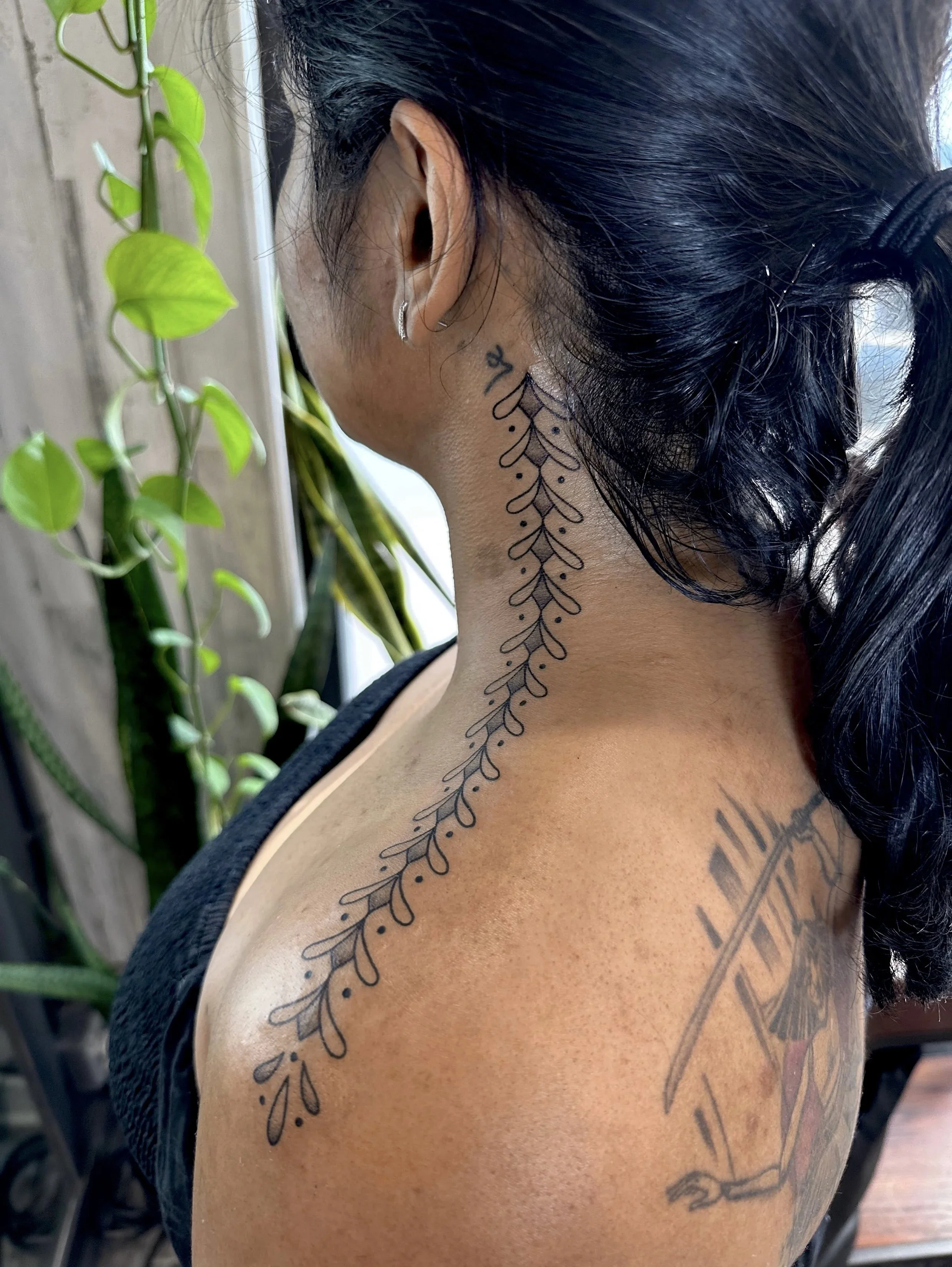A woman with black hair in a ponytail, wearing earrings, has a tattoo of a leafy vine on her neck and shoulder, against a background with green plants and a wooden wall.