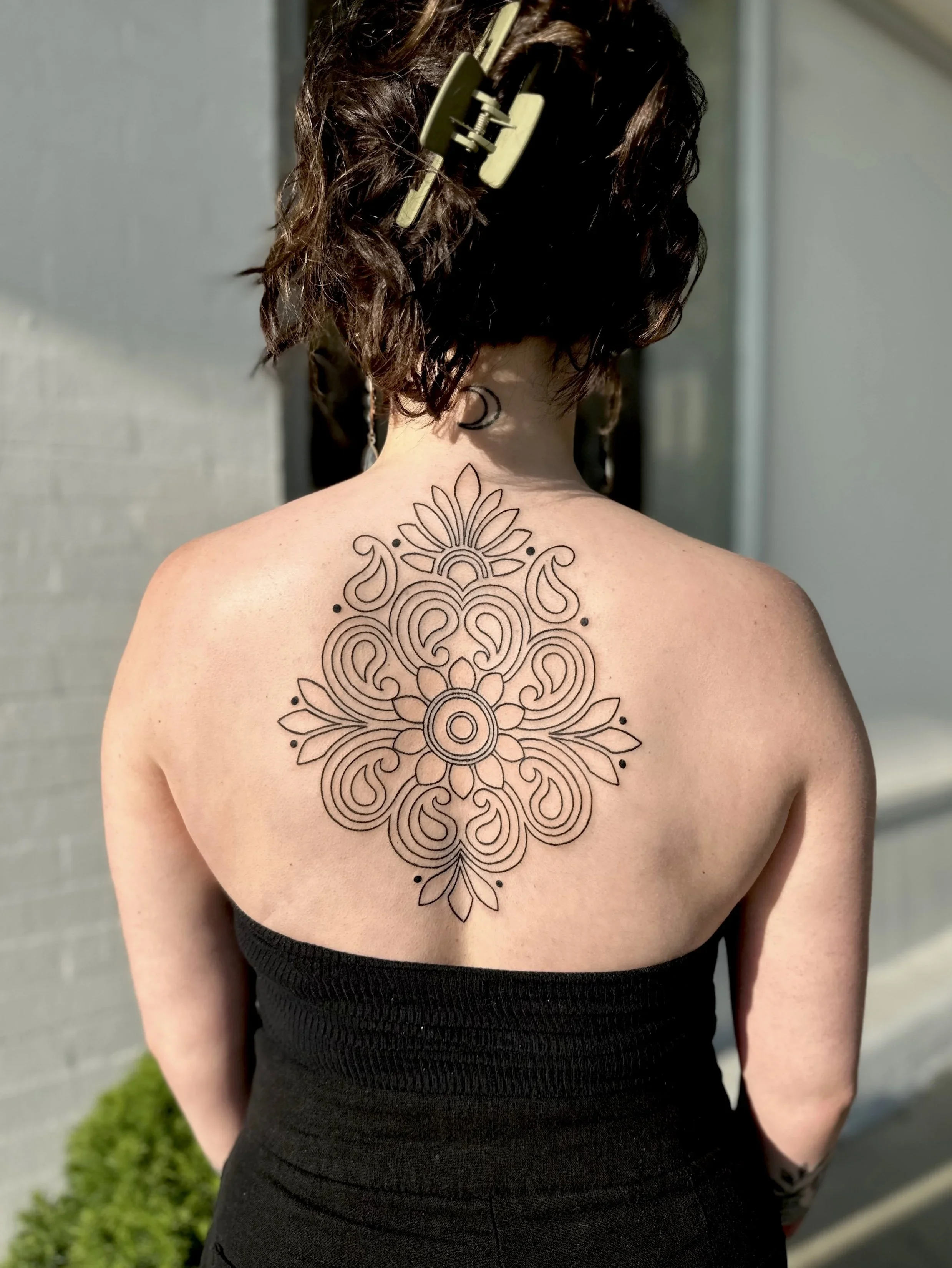 Back view of a person with short, wavy hair, wearing a hair clip, showing a large, intricate mandala-style tattoo on their upper back and shoulders.