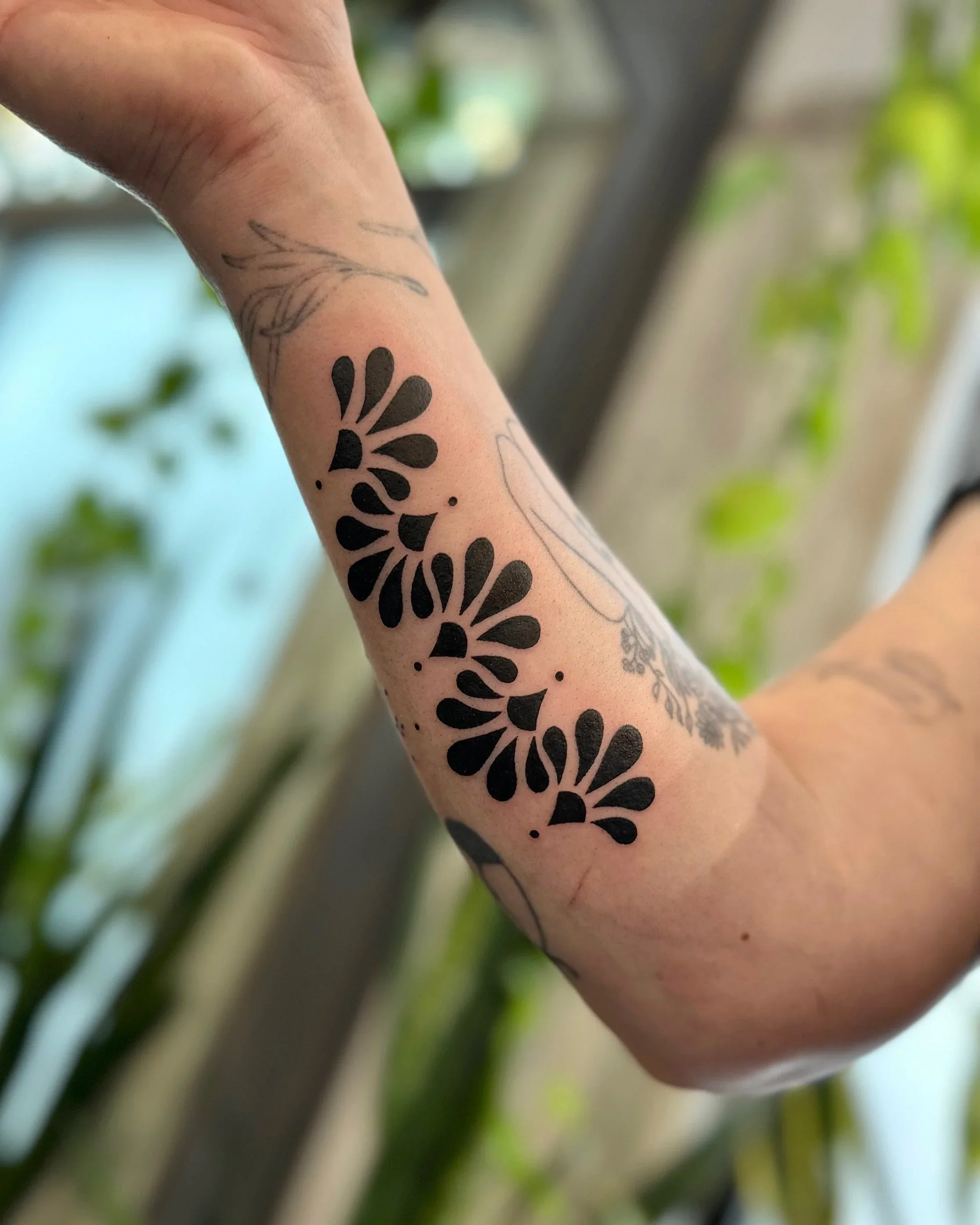Close-up of a person's arm with black floral tattoos, with a blurred green and white background.
