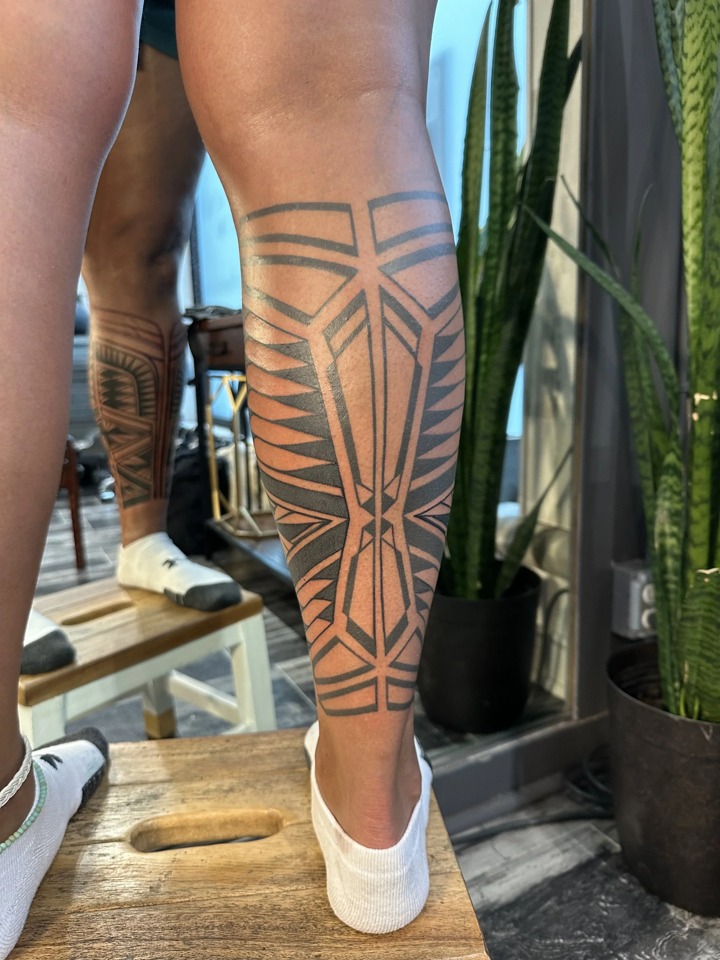 Close-up of a person's lower leg with tattoo, wearing a white sock, standing on a wooden surface, with potted plants and furniture in the background.