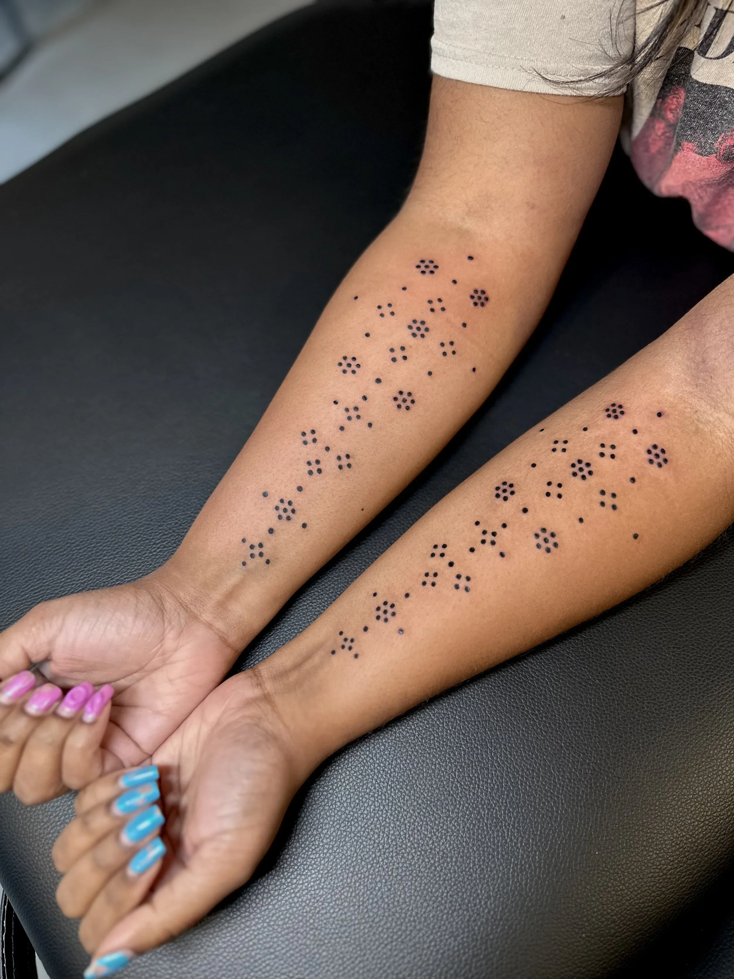 Close-up of person's arms lying on a black surface, with temporary black henna tattoo designs in floral patterns on their forearms. The person has multi-colored painted nails.