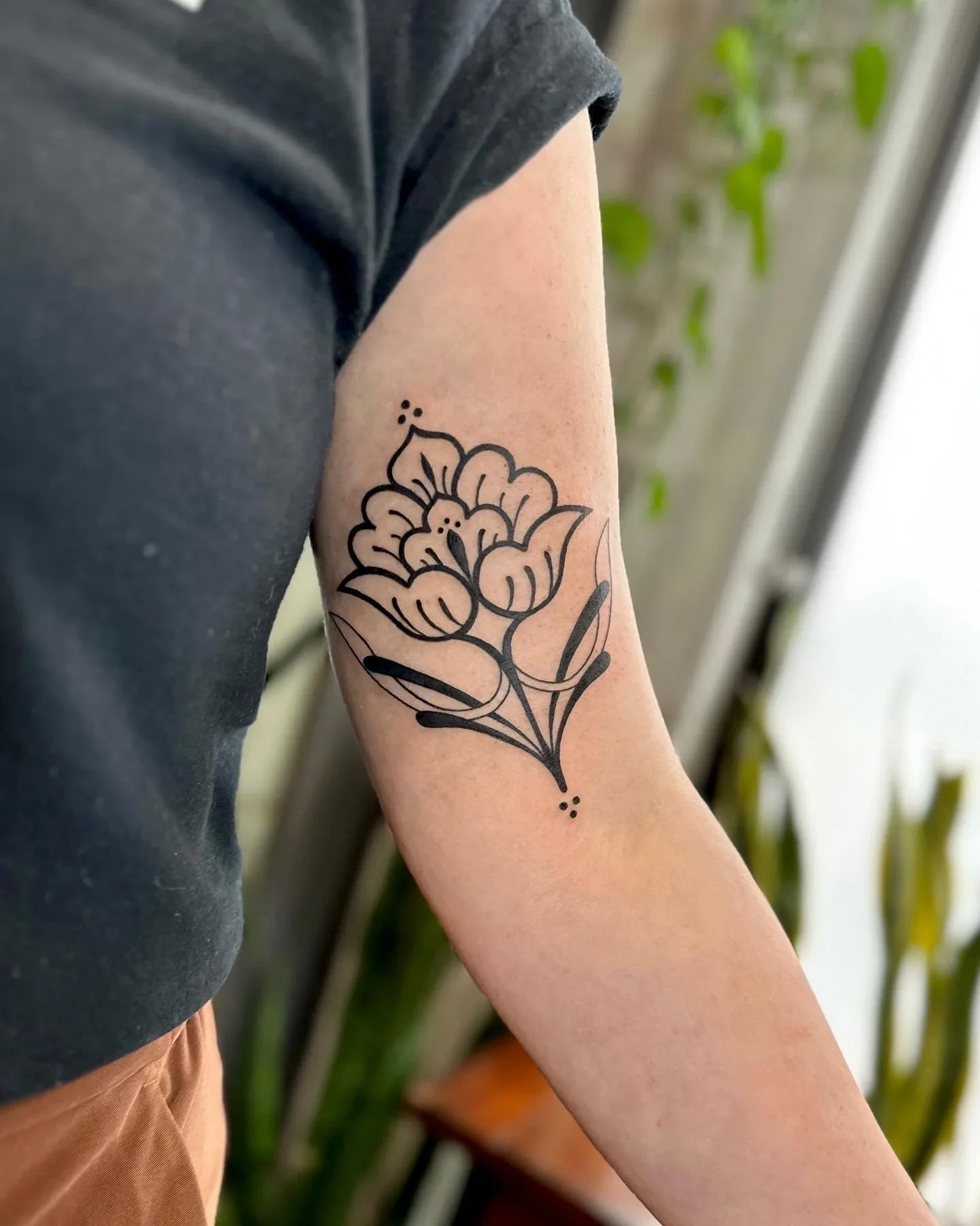 A person with a floral tattoo on their upper arm, wearing a dark t-shirt, standing indoors near green plants.