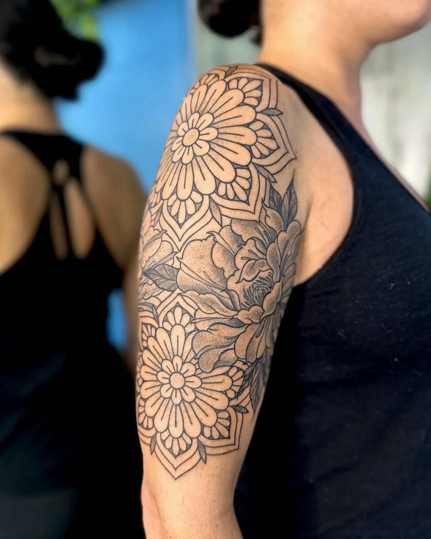 Close-up of a person's upper arm with a large black outline floral tattoo, featuring detailed petals and leaves, and a portion of their black sleeveless shirt visible.