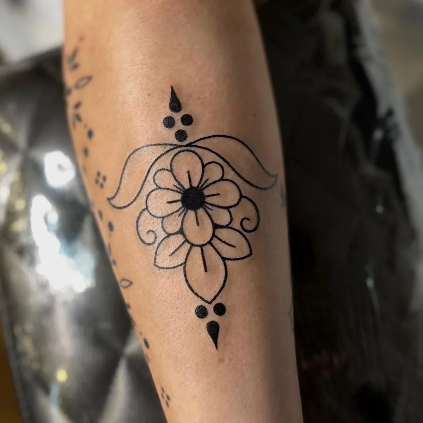 Black ink floral tattoo on skin, featuring a large flower with five petals and swirl details, connected with dotted lines and decorative elements.