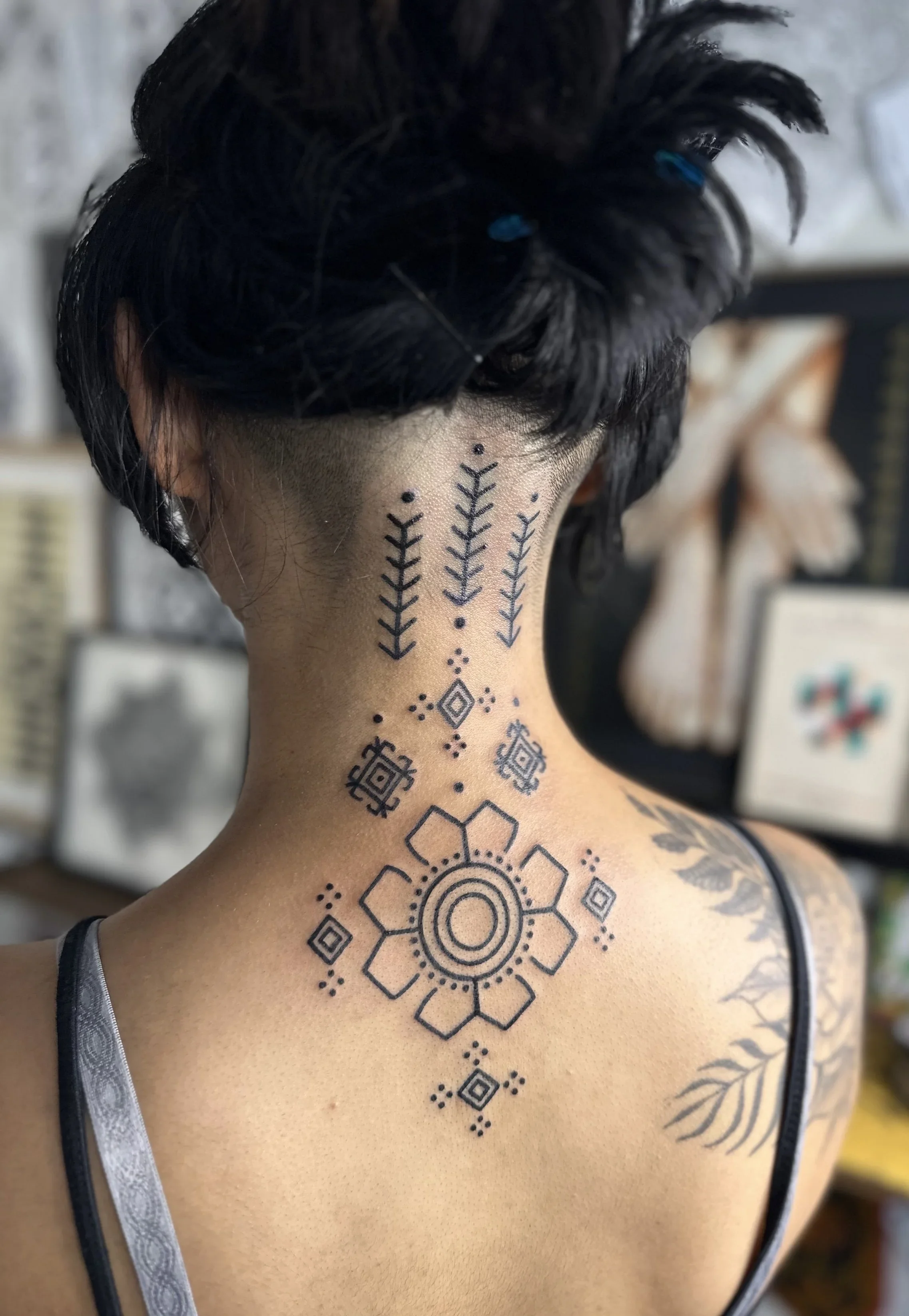 Back of a person's neck and shoulder showing a geometric tattoo design extending onto the shoulder, with part of a black strap visible on the person's shoulder.