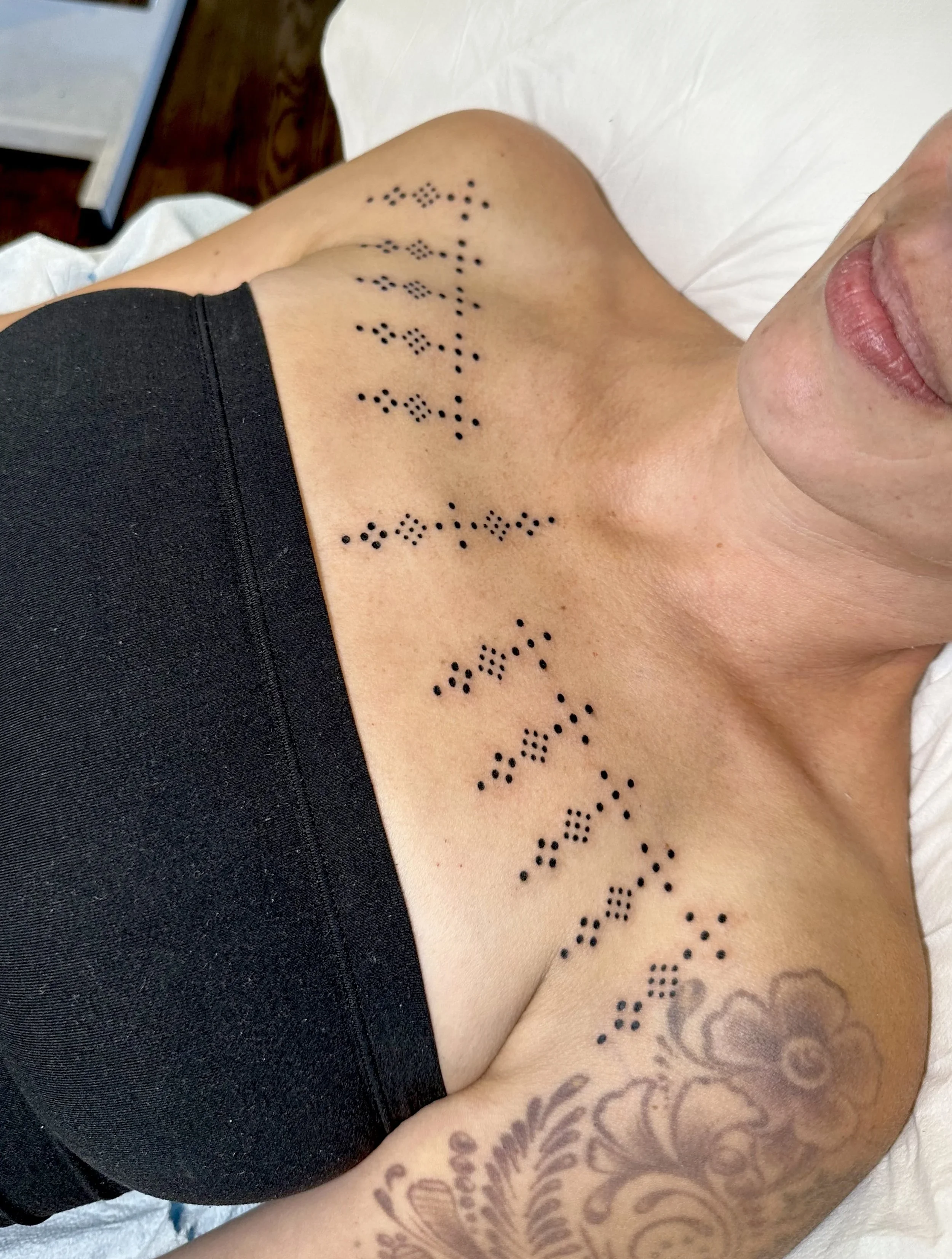 Close-up photo of a person's upper arm and shoulder, showing intricate black tattoo designs including geometric patterns and floral elements, with part of their face and lips visible.