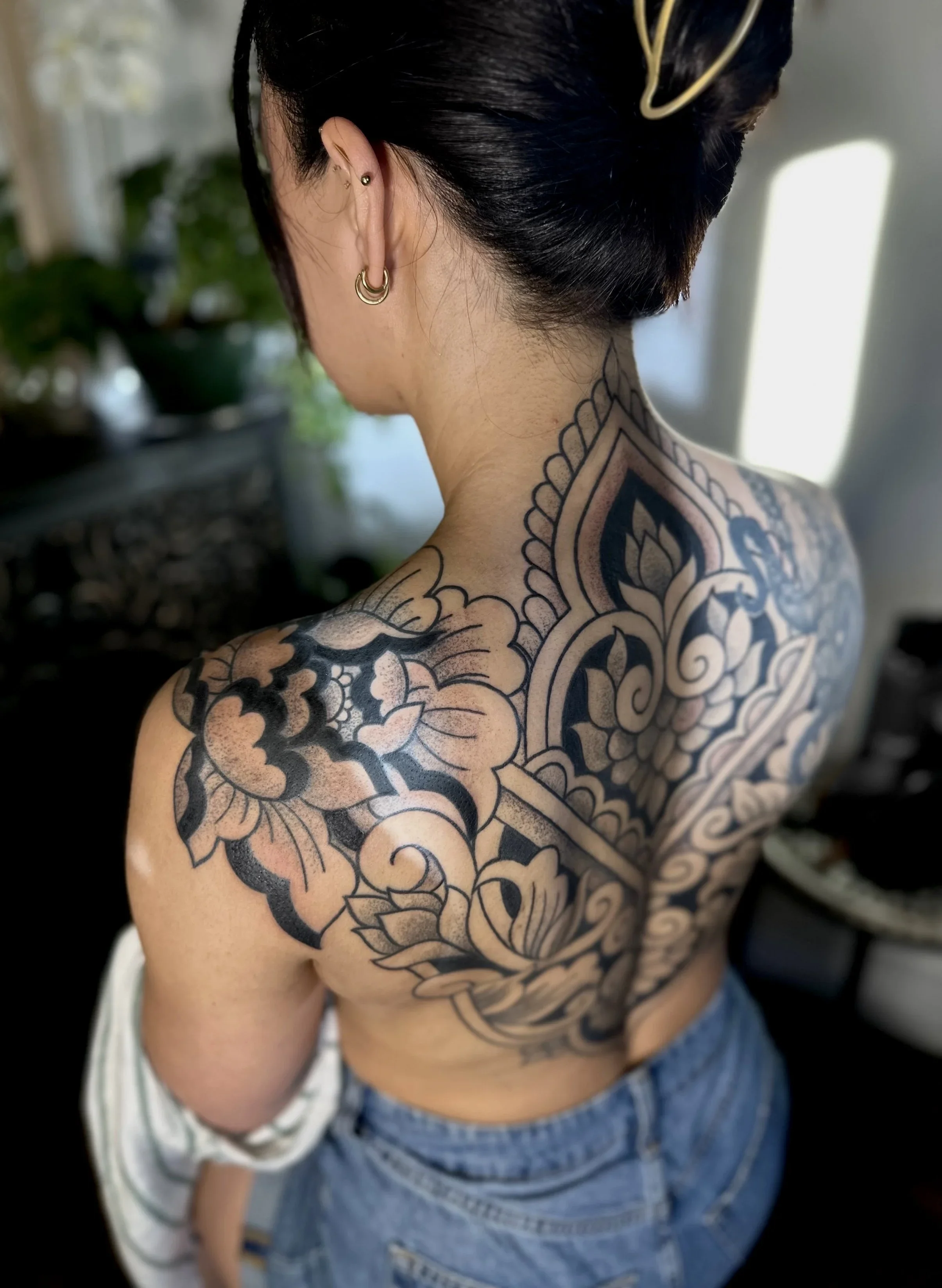 Back view of a woman with tattoos on her shoulder and neck, wearing gold earrings, and with dark hair tied up with a hair clip.