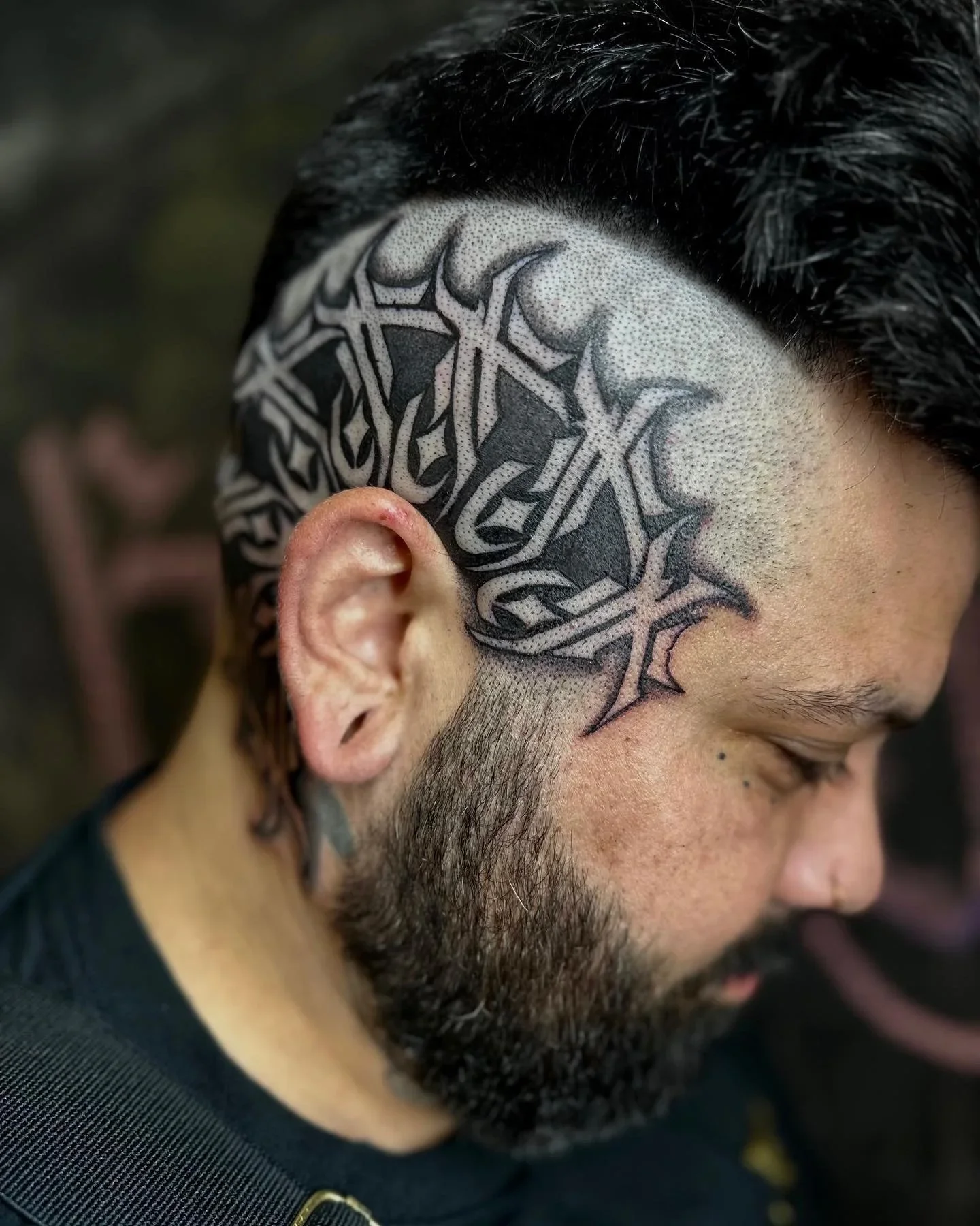 Close-up of a man's head showing a detailed black and grey tattoo behind his ear and side of the head, with short dark hair and a beard.
