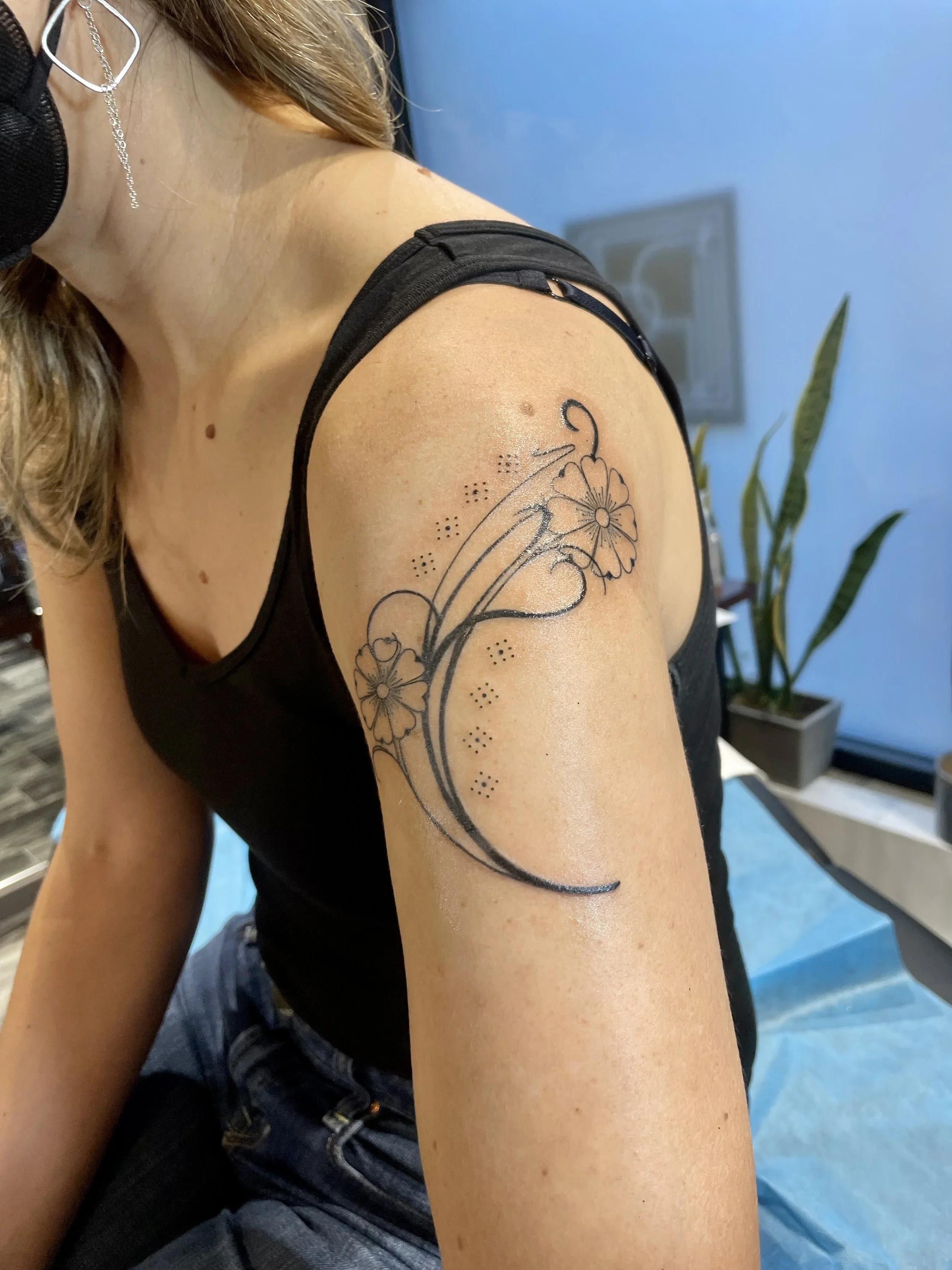 A woman with a floral moon tattoo on her upper arm, wearing a black tank top and a face mask, sitting indoors near a plant with long leaves.