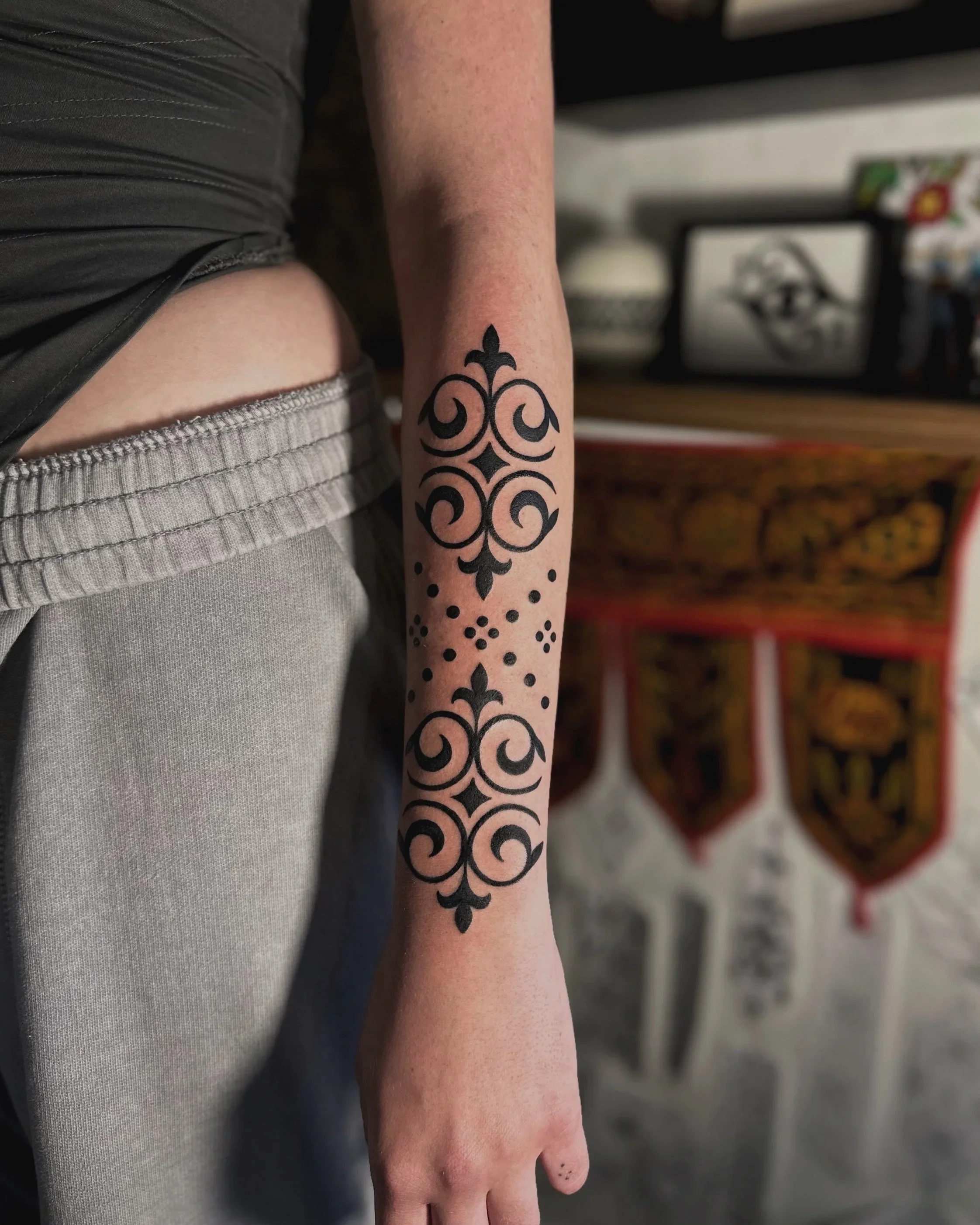 Close-up of a person's arm and hand, displaying a black tribal-style tattoo on the forearm with intricate swirl and dot patterns, in an indoor setting.