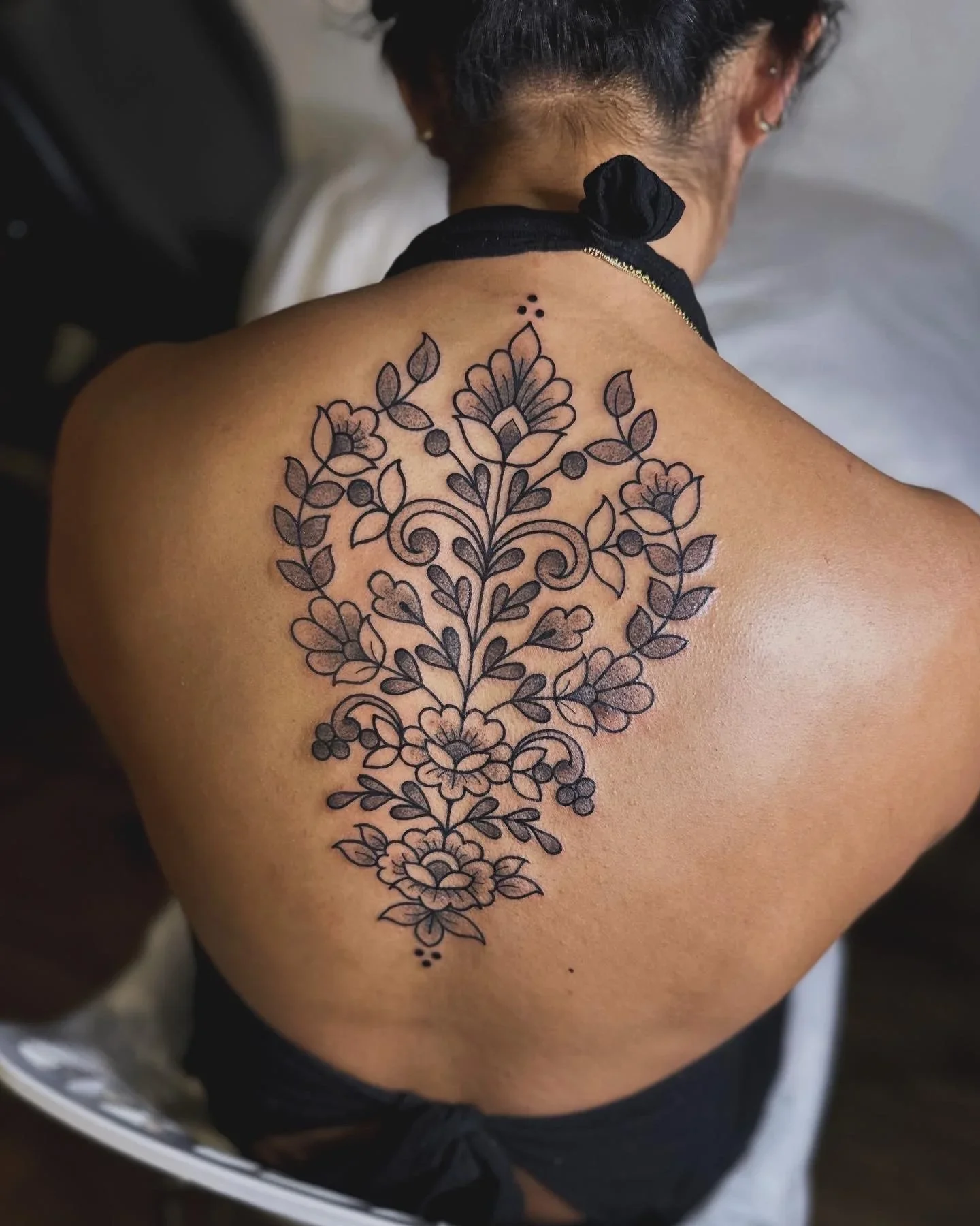 Back of a person with a floral tattoo on their upper back and shoulder, featuring intricate outlines of flowers and leaves.