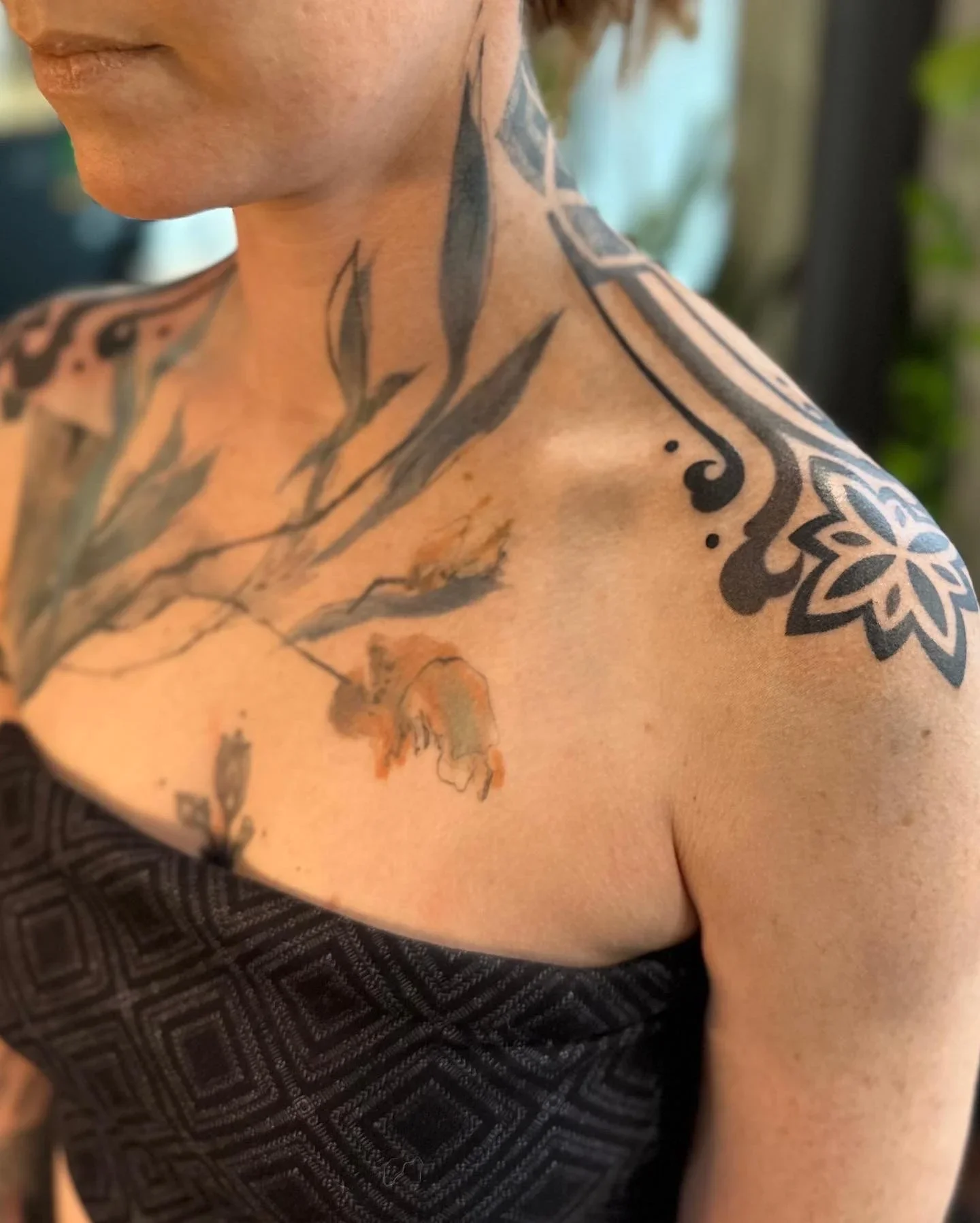 Close-up of a person's shoulder and neck with tattoos, including a floral design on the shoulder and a branch with leaves on the neck, wearing a black patterned top.