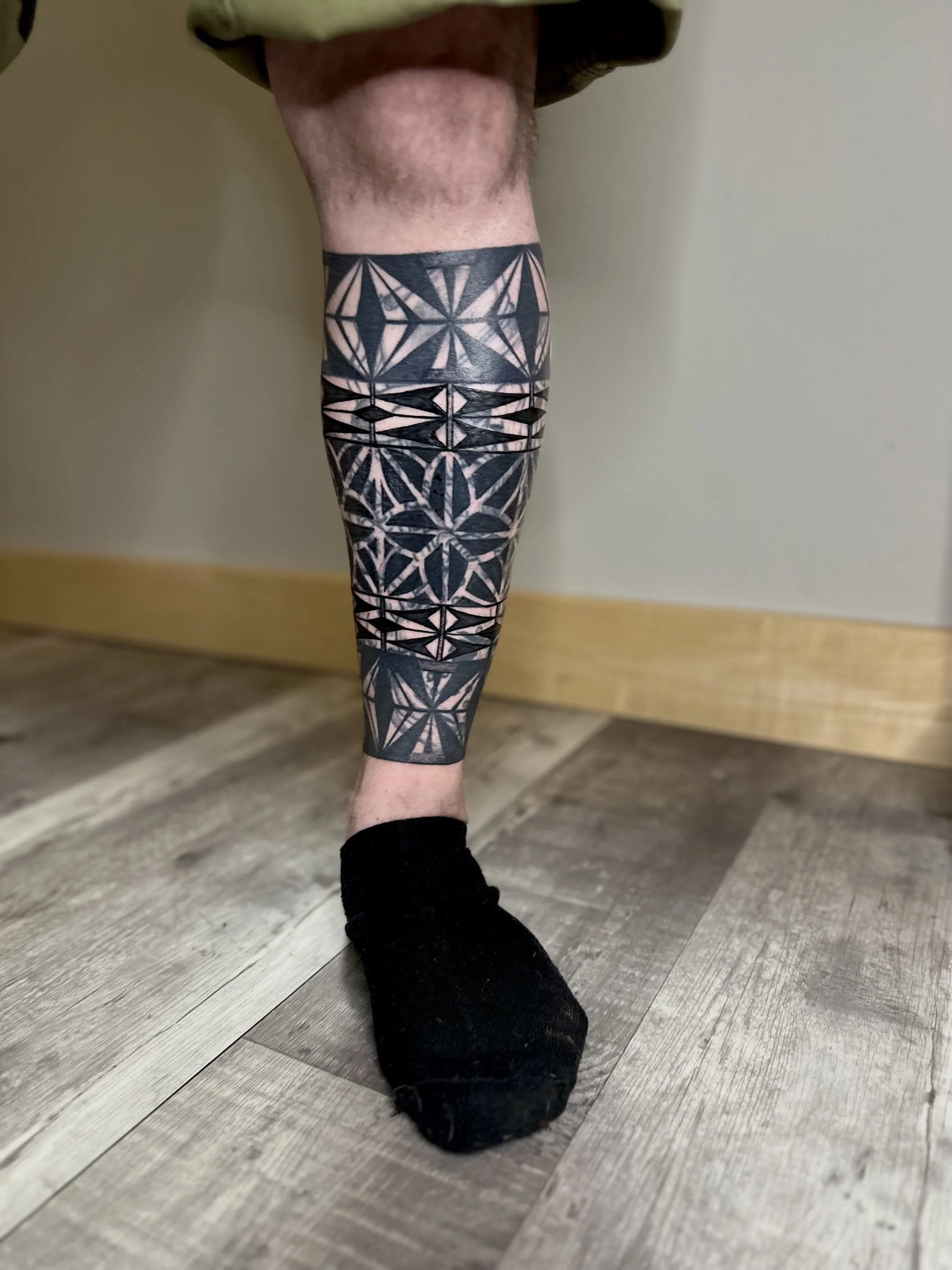 A person's lower leg and foot wearing a patterned tattoo resembling geometric shapes in black ink, with the person standing on a wooden floor, wearing a black sock on one foot.