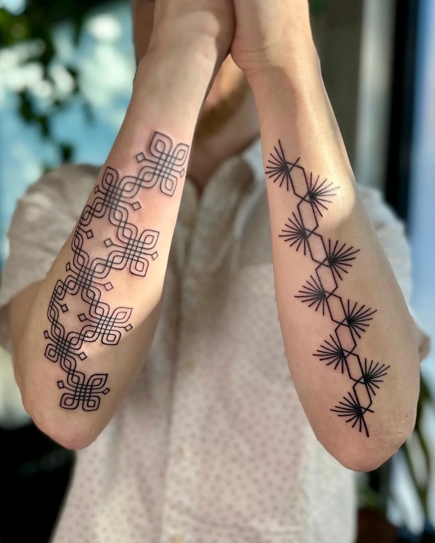 Two arms with geometric Tattoos, one on each arm. The tattoo on the left arm features intricate interwoven lines, and the tattoo on the right arm features polygonal shapes with radiating lines.