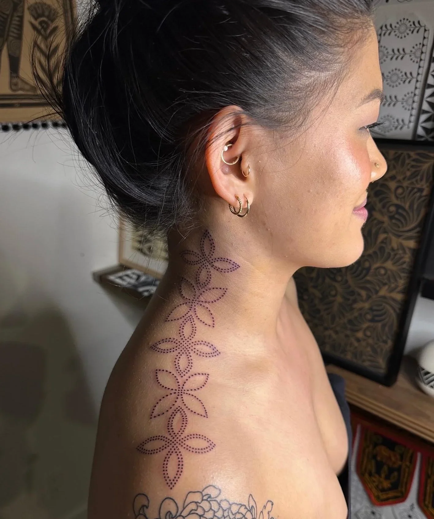Side profile of a woman with black hair styled back, wearing multiple earrings, and having tattoos on her neck and shoulder. The tattoos include a leaf pattern on her neck and floral design on her shoulder.