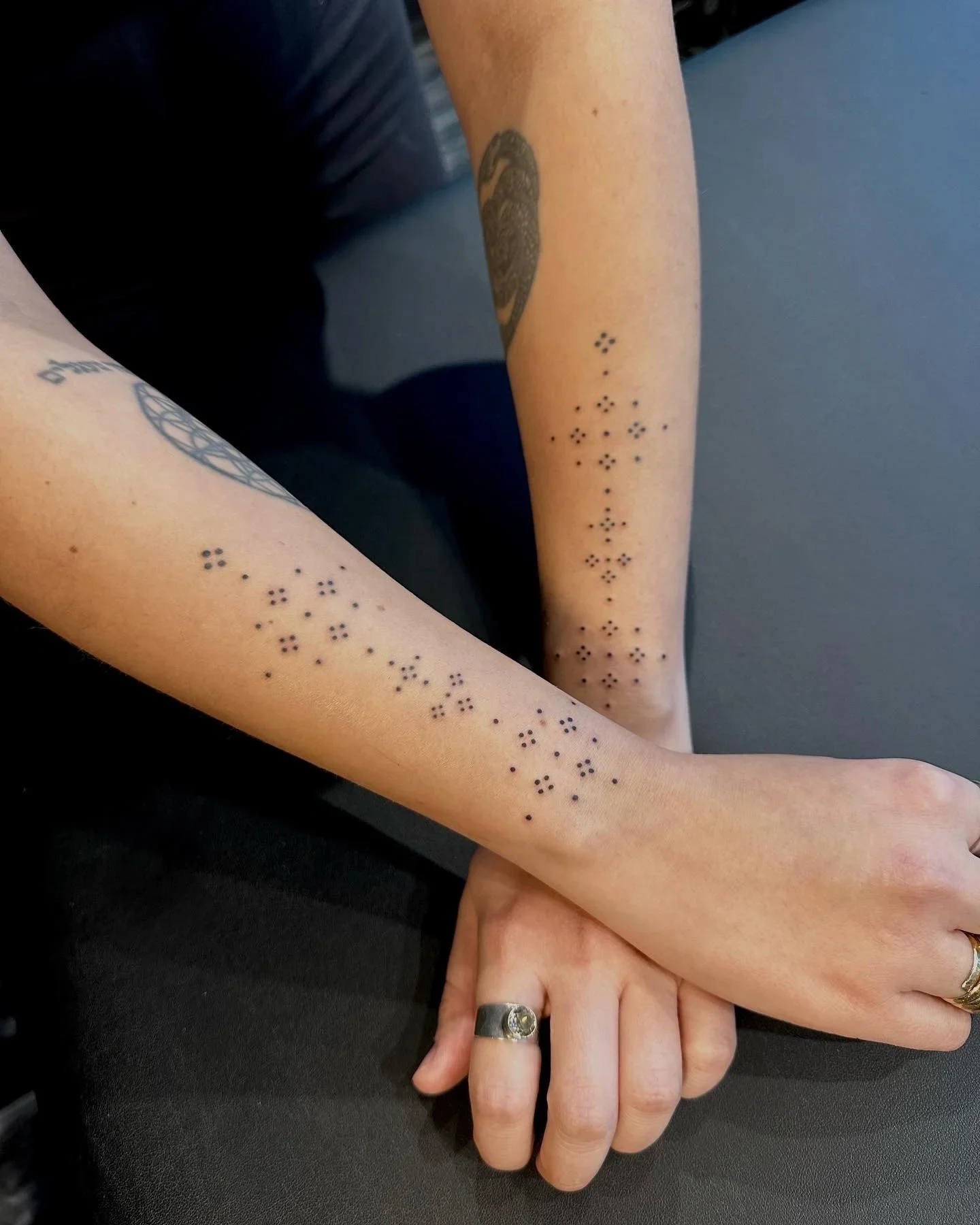 Two people holding hands, one with a tattoo of a swan on their arm and small dot tattoos on the other arm, sitting on a dark surface.