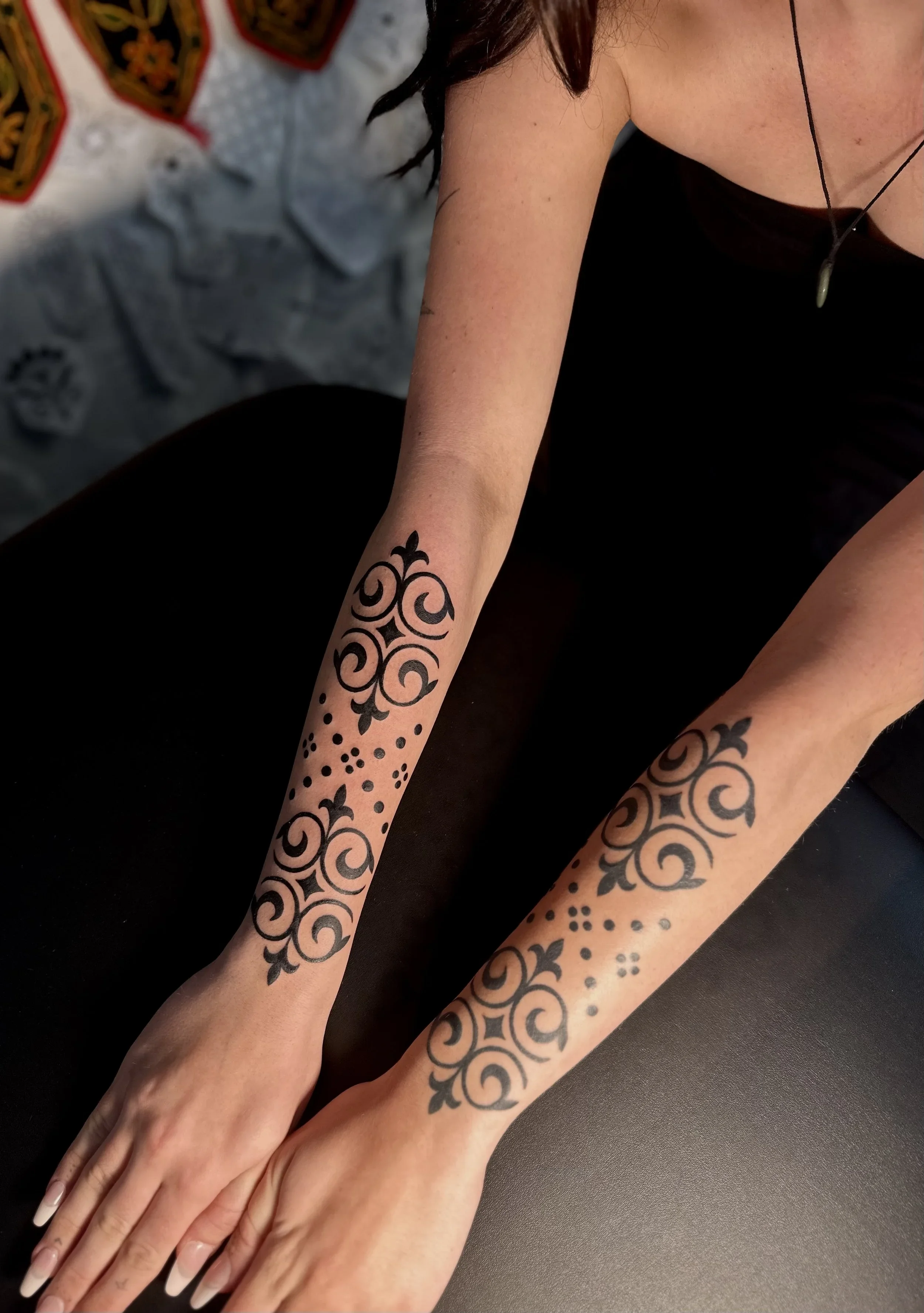 A person's arms and hands are resting on a black surface, displaying a tattoo sleeve with black, intricate, tribal or ornamental designs on the forearms.