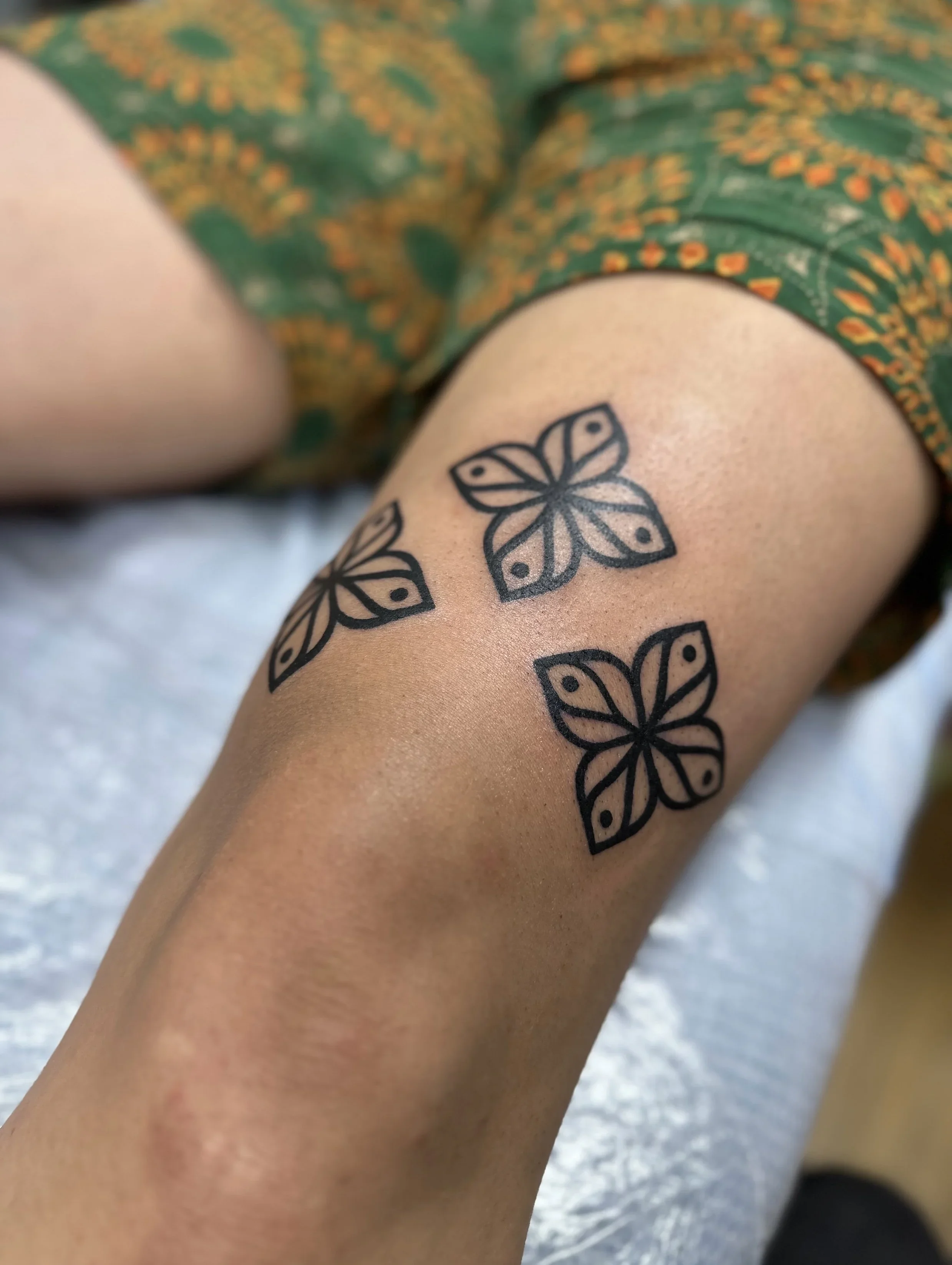 Close-up of a person's upper arm with a tattoo of a geometric butterfly design, along with part of a green and yellow patterned garment.