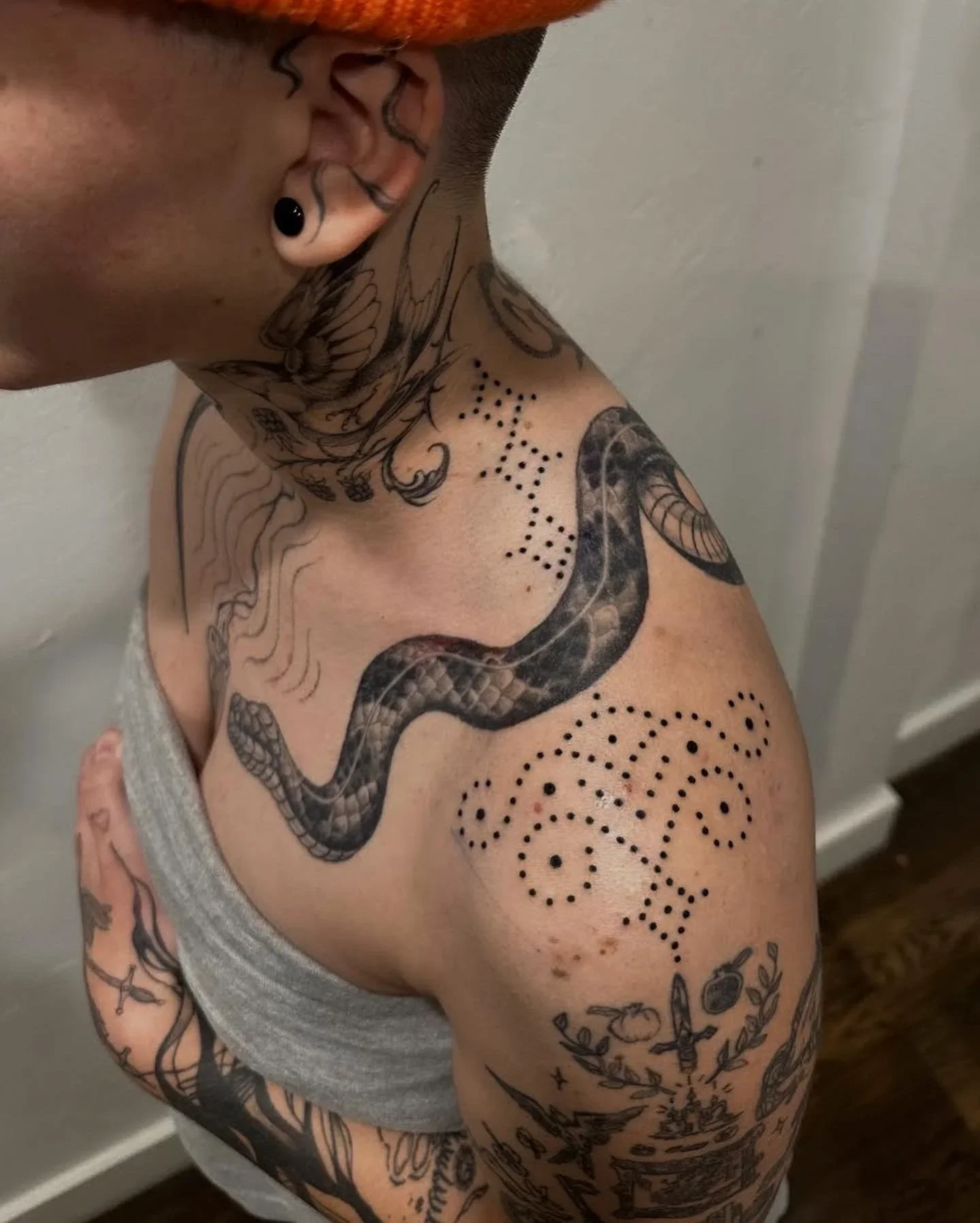 A person with tattoos on their neck, chest, and arms, showing a snake tattoo on their shoulder and various other designs, including dots and floral patterns.