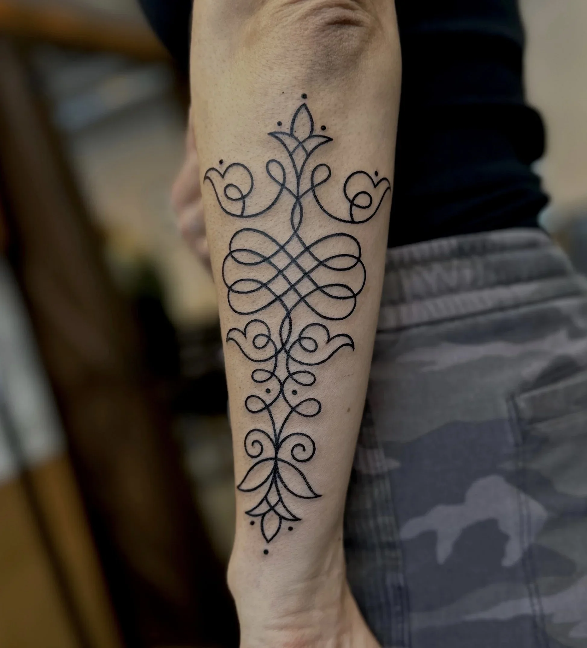 A person's forearm with a black line tattoo of a floral design and symmetrical scrollwork.