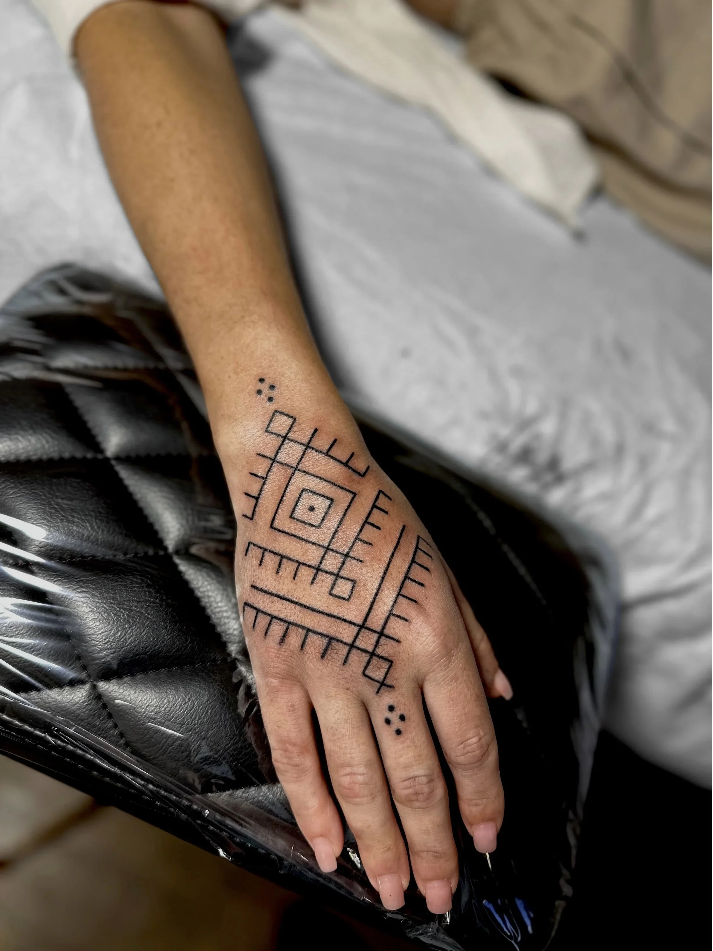 A human hand with geometric tattoo design on the wrist, resting on a black leather surface. The tattoo includes various lines, squares, and dots.