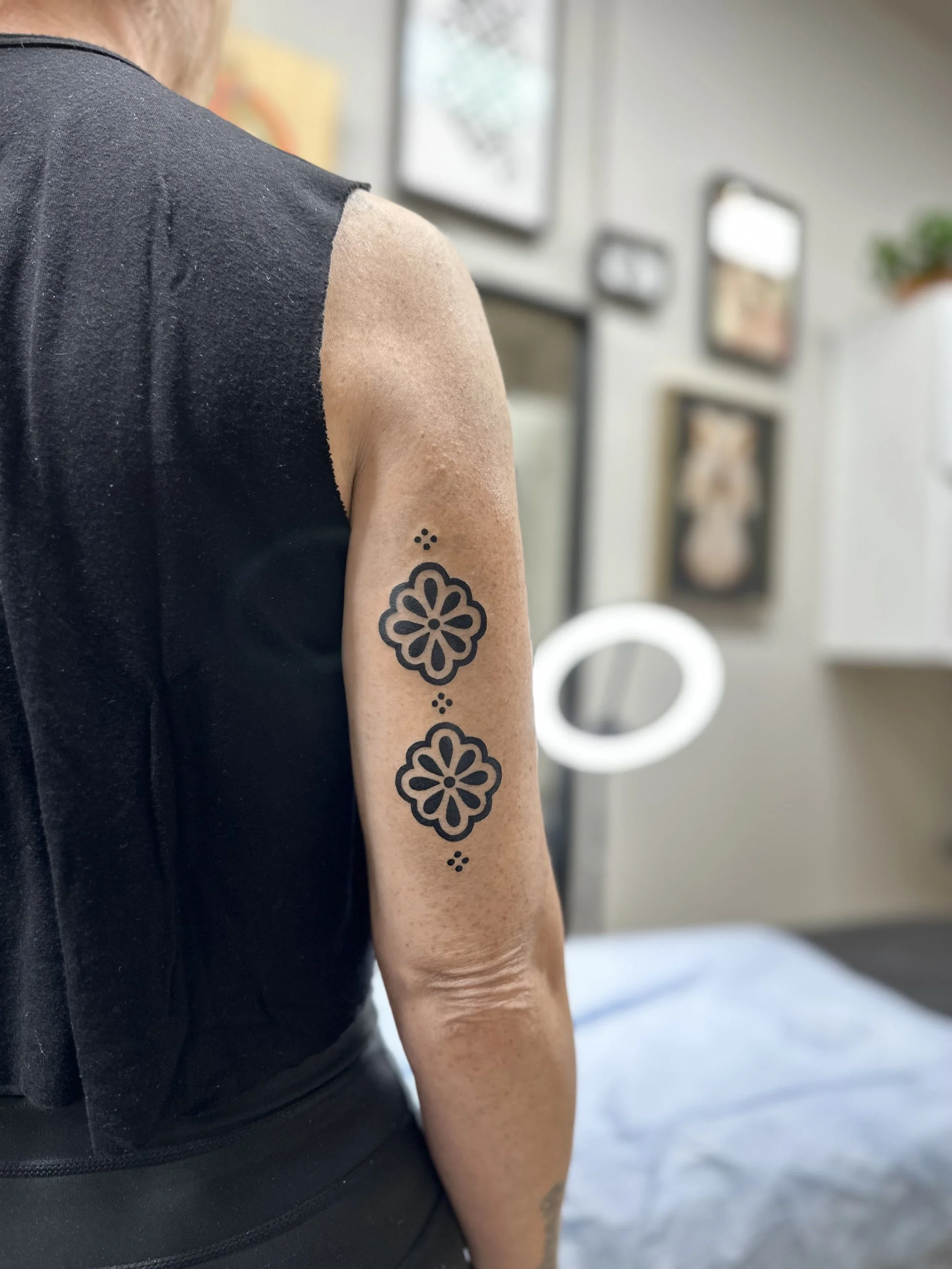 Close-up of a person's right arm with a black floral tattoo and a sleeveless black shirt, in a room with framed pictures on the wall and a window in the background.