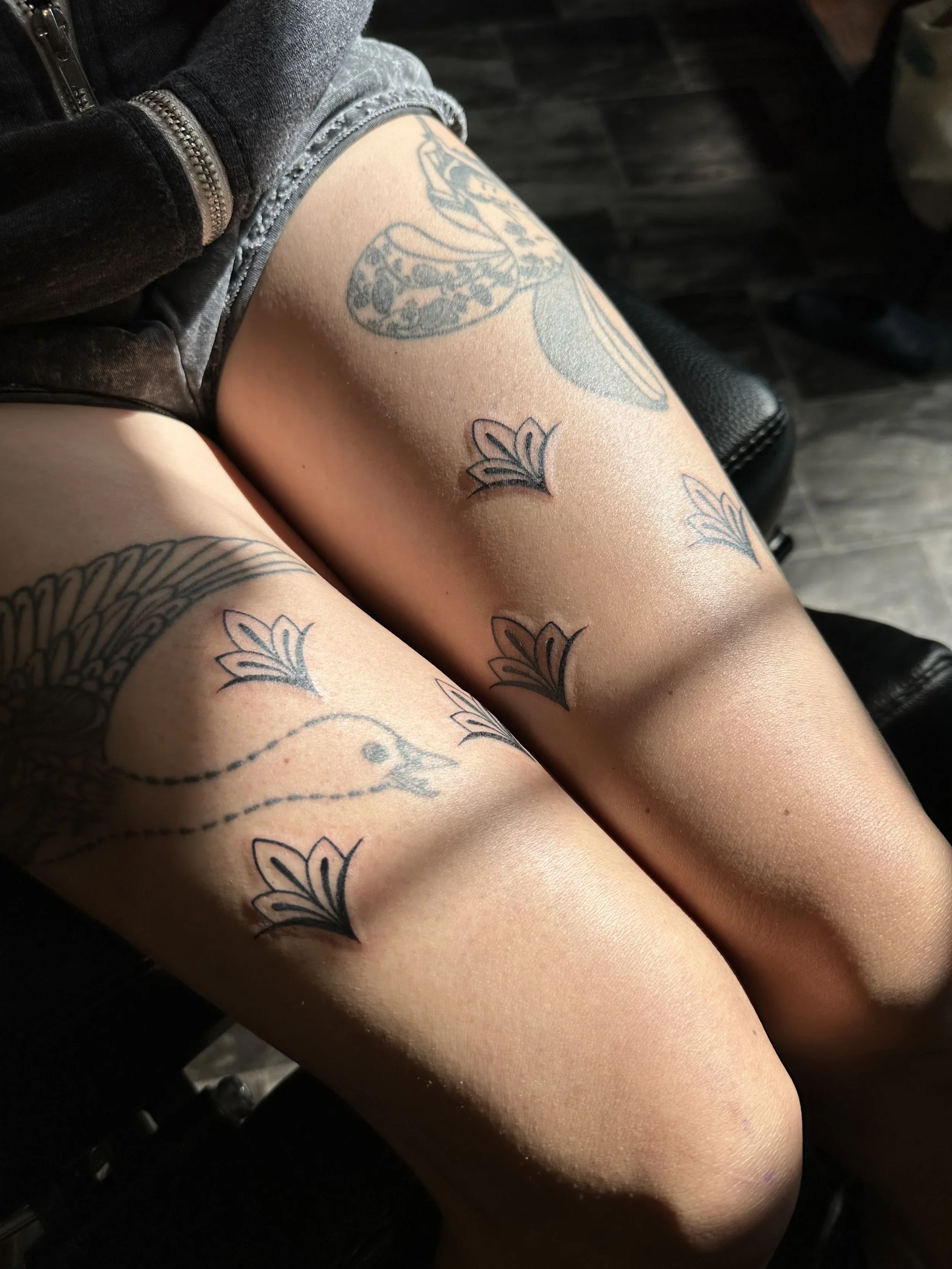 Close-up of a person's upper thighs with multiple black outline tattoos of butterflies and flowers, with some shading, against a background of dark wood flooring.