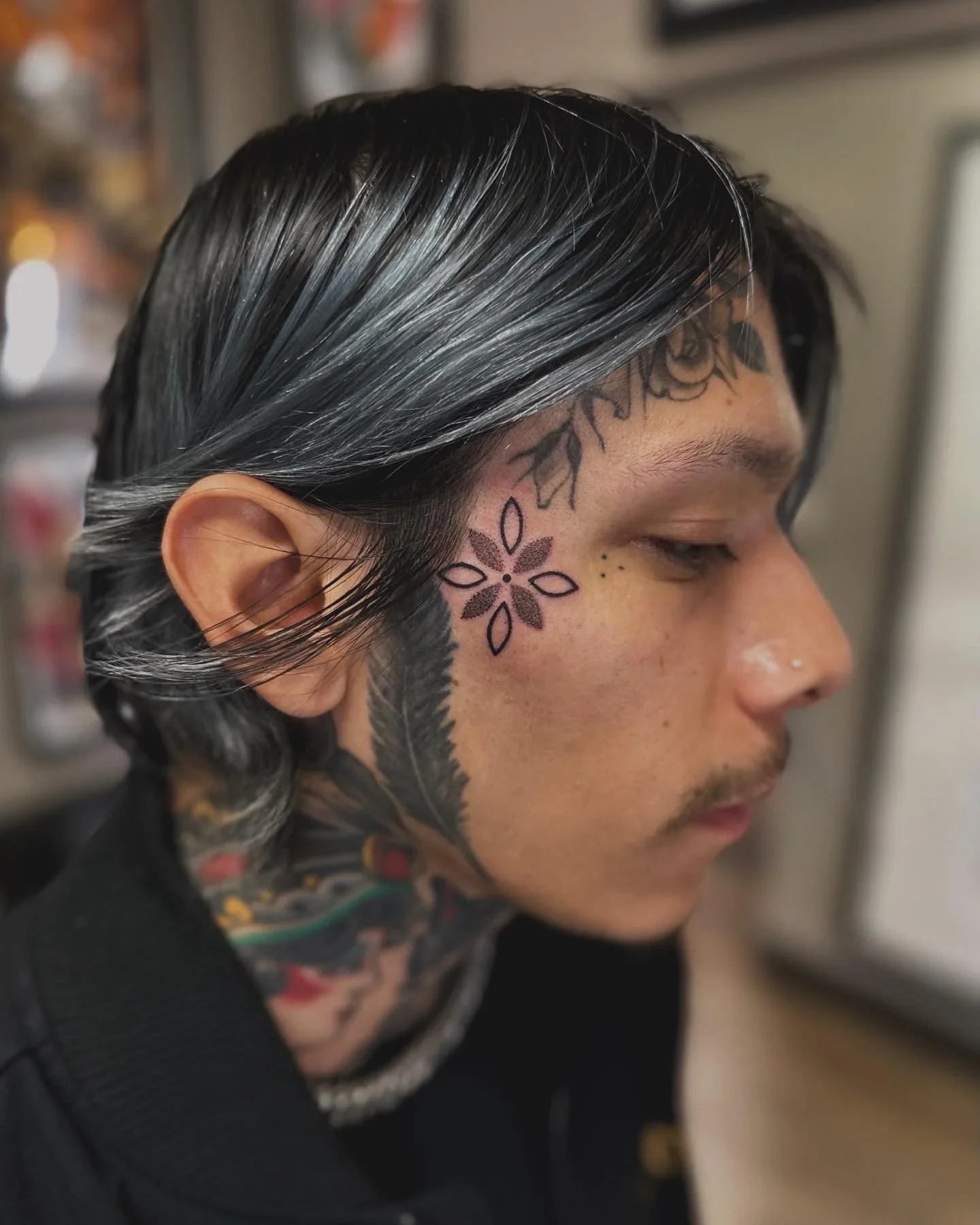 Close-up side profile of a person with long black hair with gray streaks, tattoos on the face and neck including a flower and leaf design.