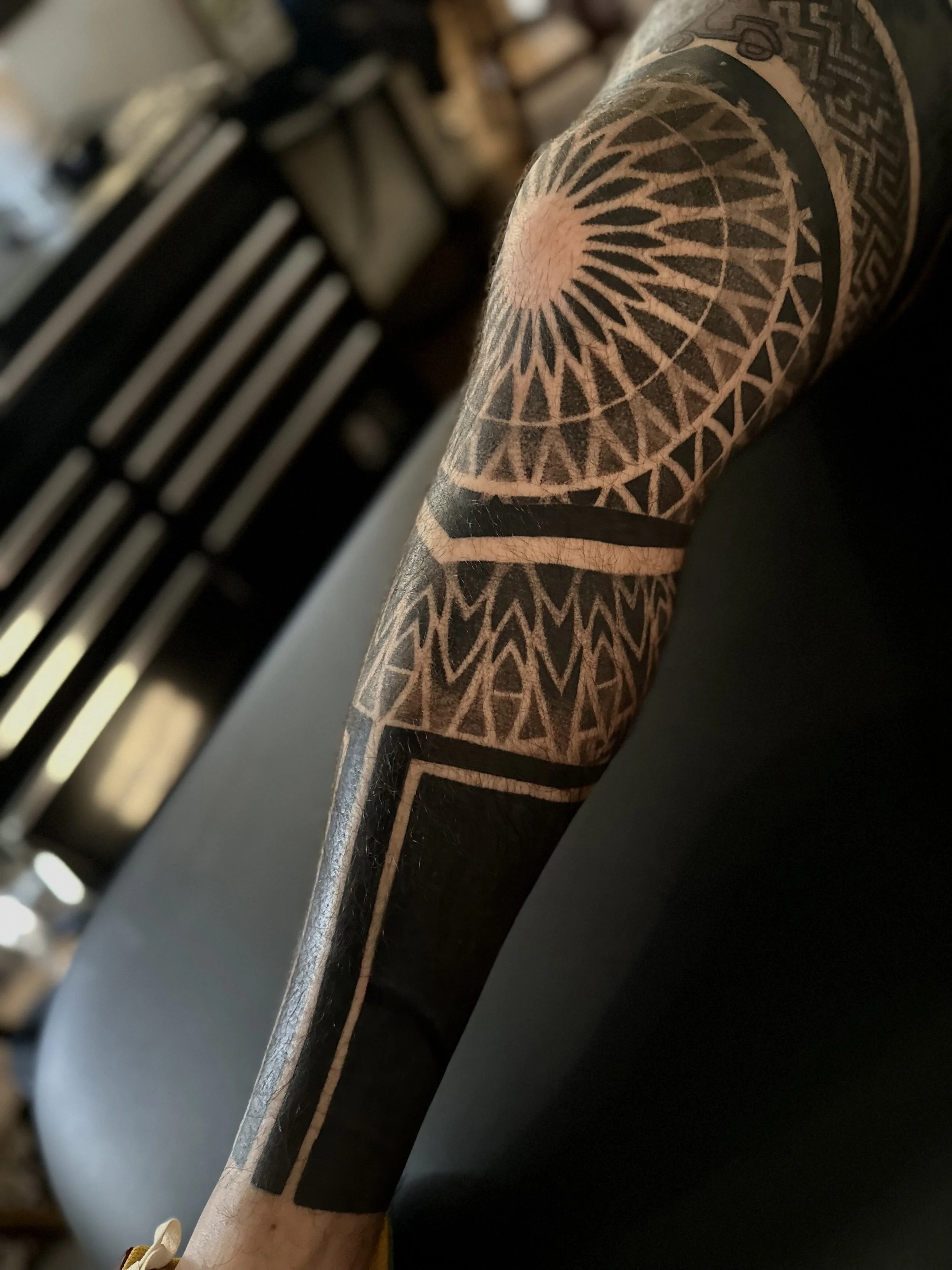 A close-up of a tattooed arm with geometric black ink designs, featuring concentric circles and triangular patterns, resting on a black surface.