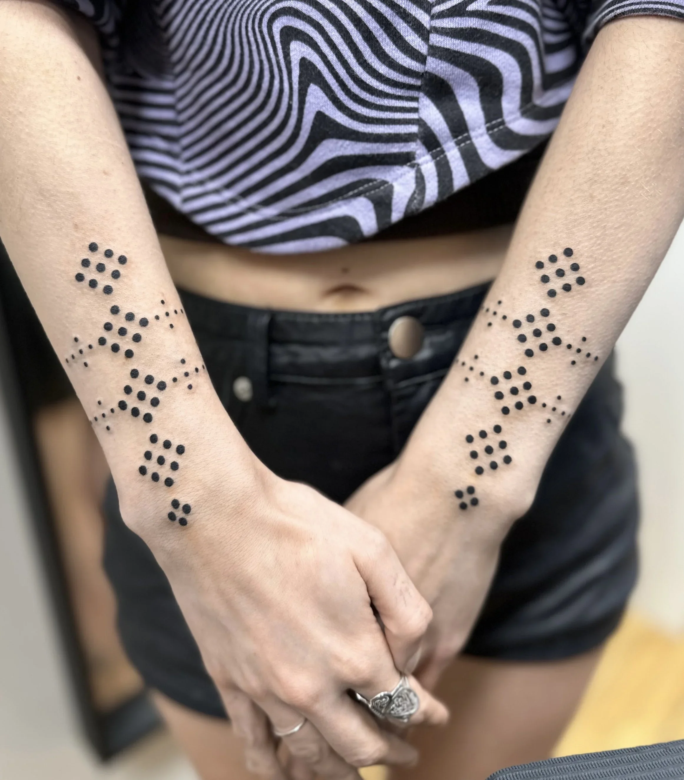 Close-up of a person's hand with multiple rings, showing tattoos of dots and lines on the arm, wearing a black and white striped shirt.