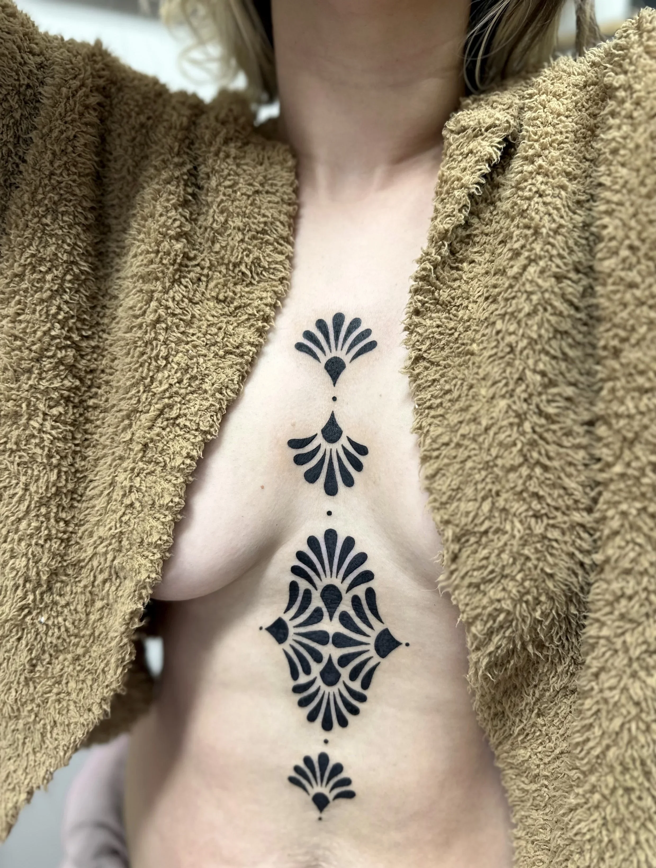 Close-up of a woman's chest with a black tattoo design running down the center, partially covered by a brown fuzzy coat.