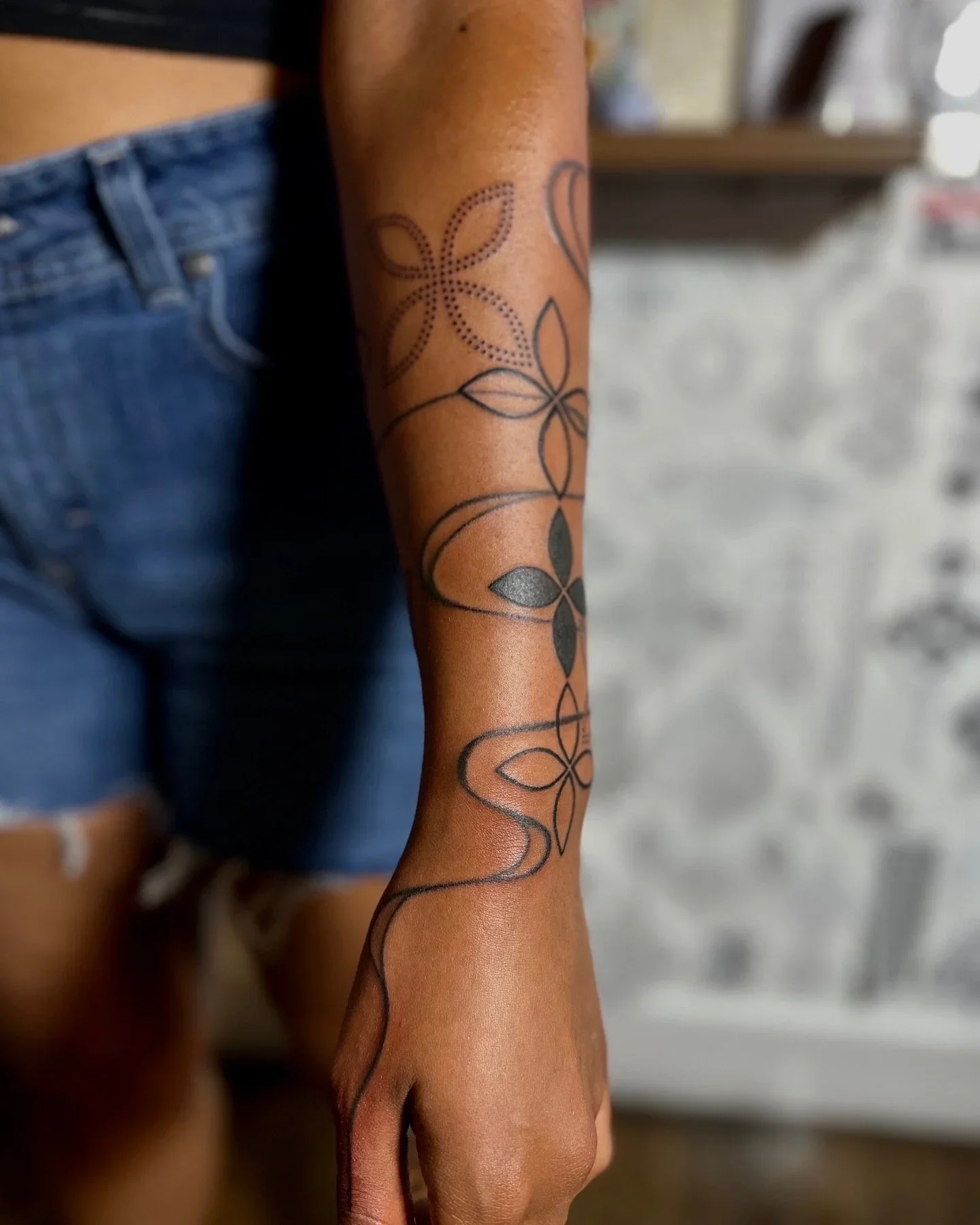A person's arm and hand with a flower-shaped tattoo pattern on the forearm, wearing blue denim shorts.