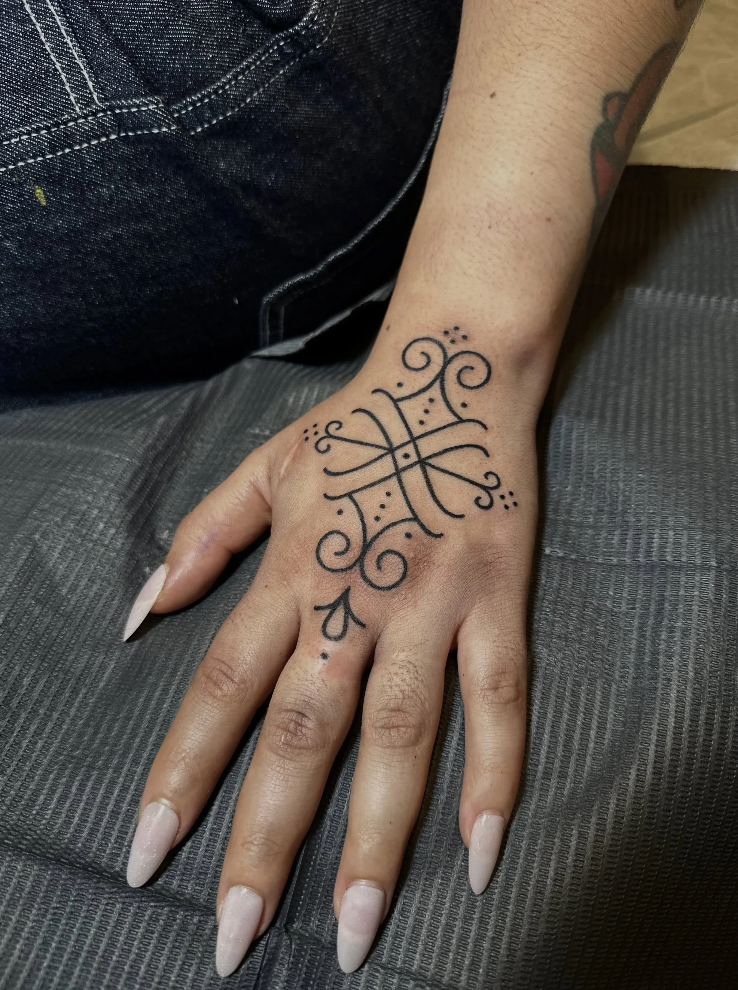 Close-up of a person's hand with long, white, almond-shaped nails. The hand has a tattoo with intricate black line designs on the wrist over the skin.