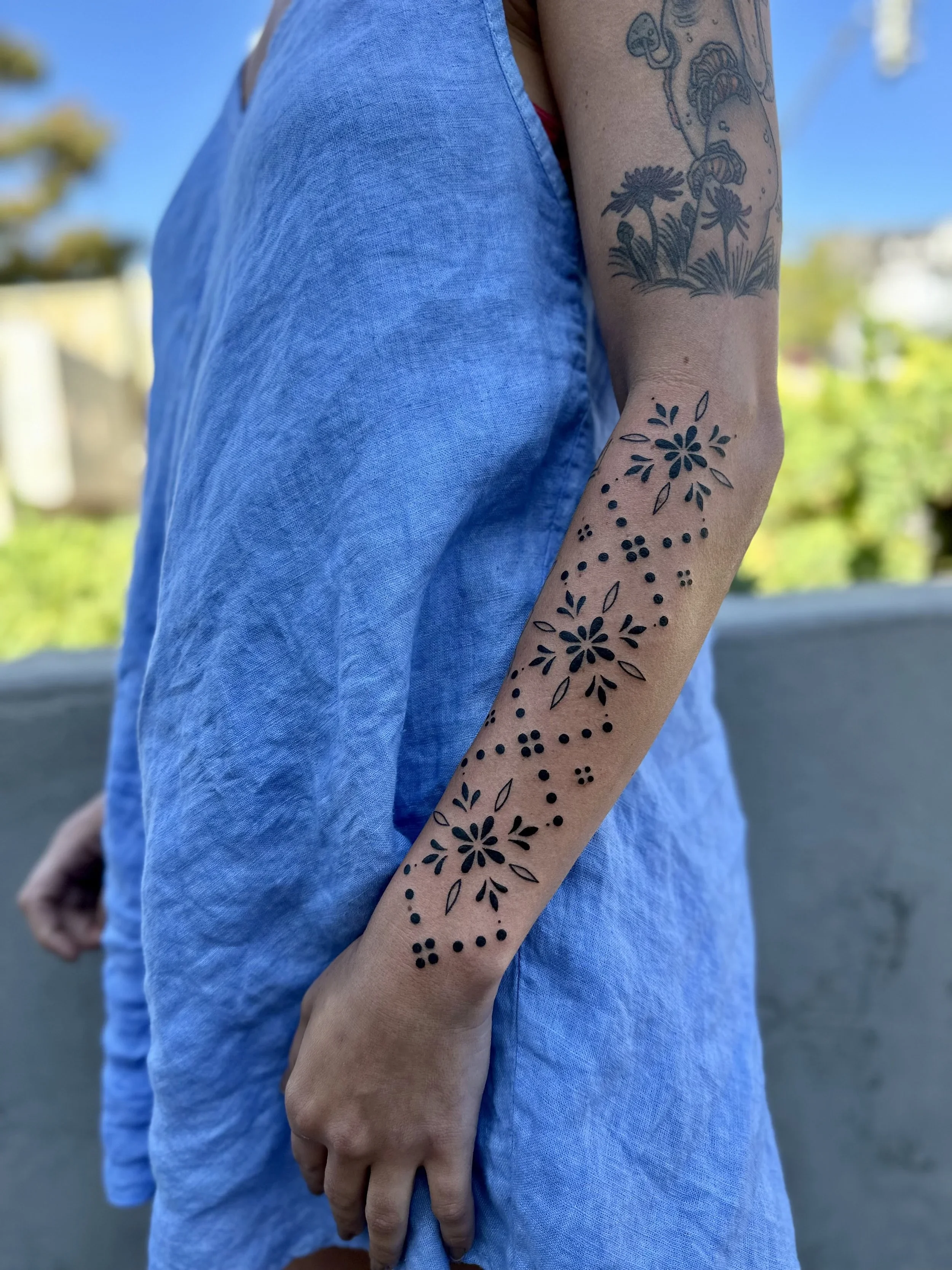 A person's arm with two tattoos, one of mushrooms and another of a floral design, visible in front of a light blue shirt and outdoors background.
