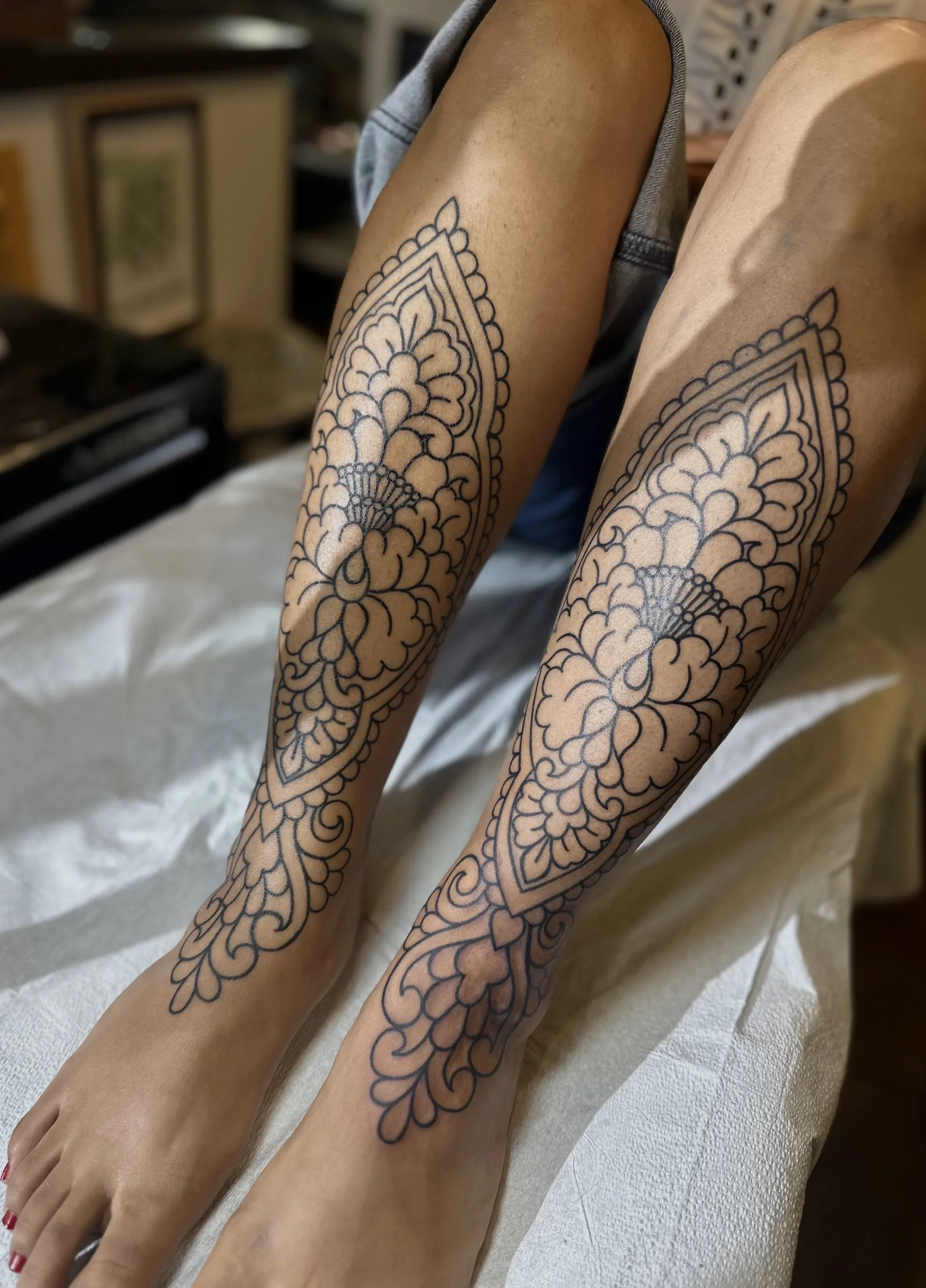 Close-up of two legs with intricate black line tattoos of floral and geometric patterns.