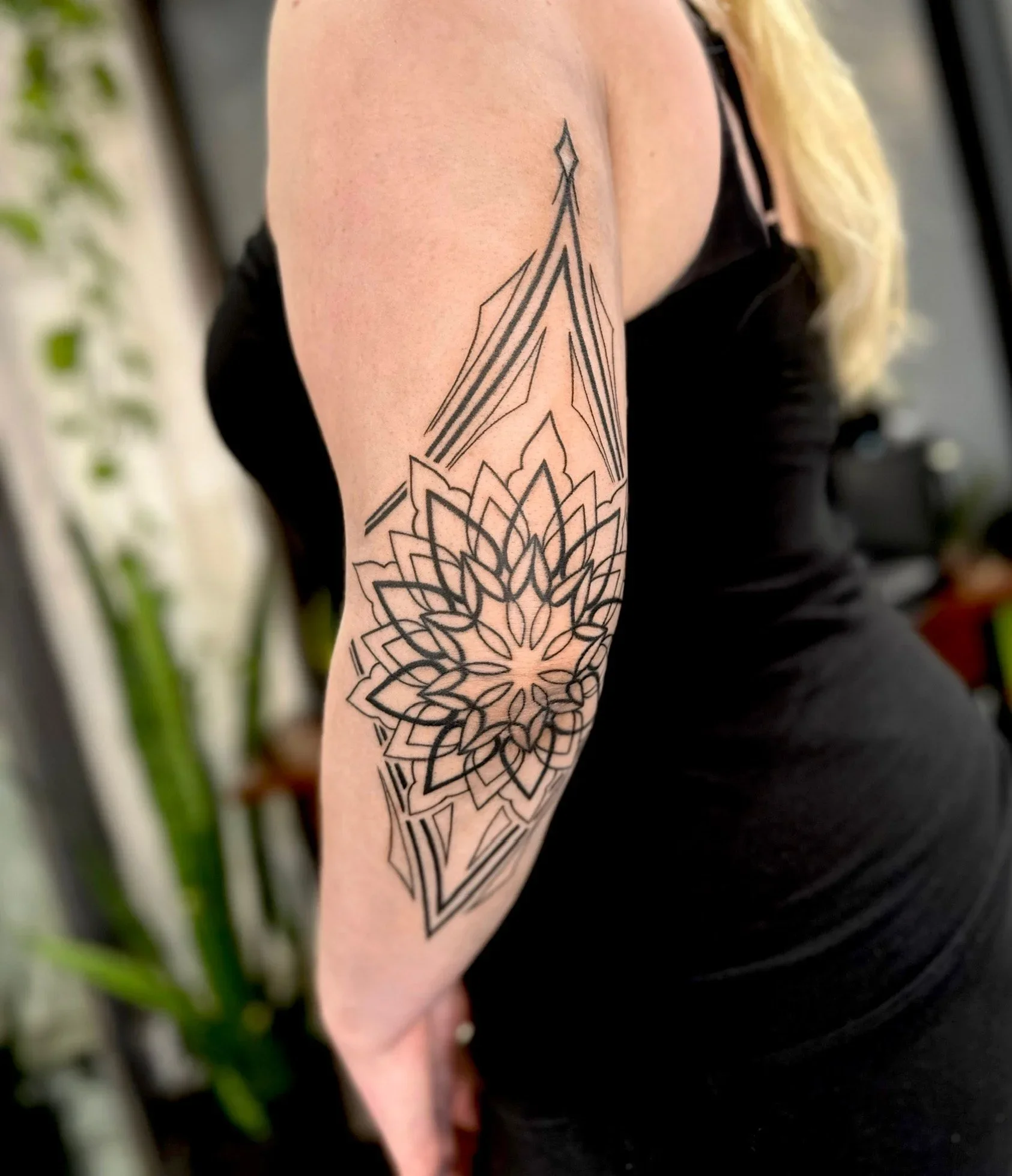 A person's upper arm with a black geometric tattoo that resembles a mandala or floral design.