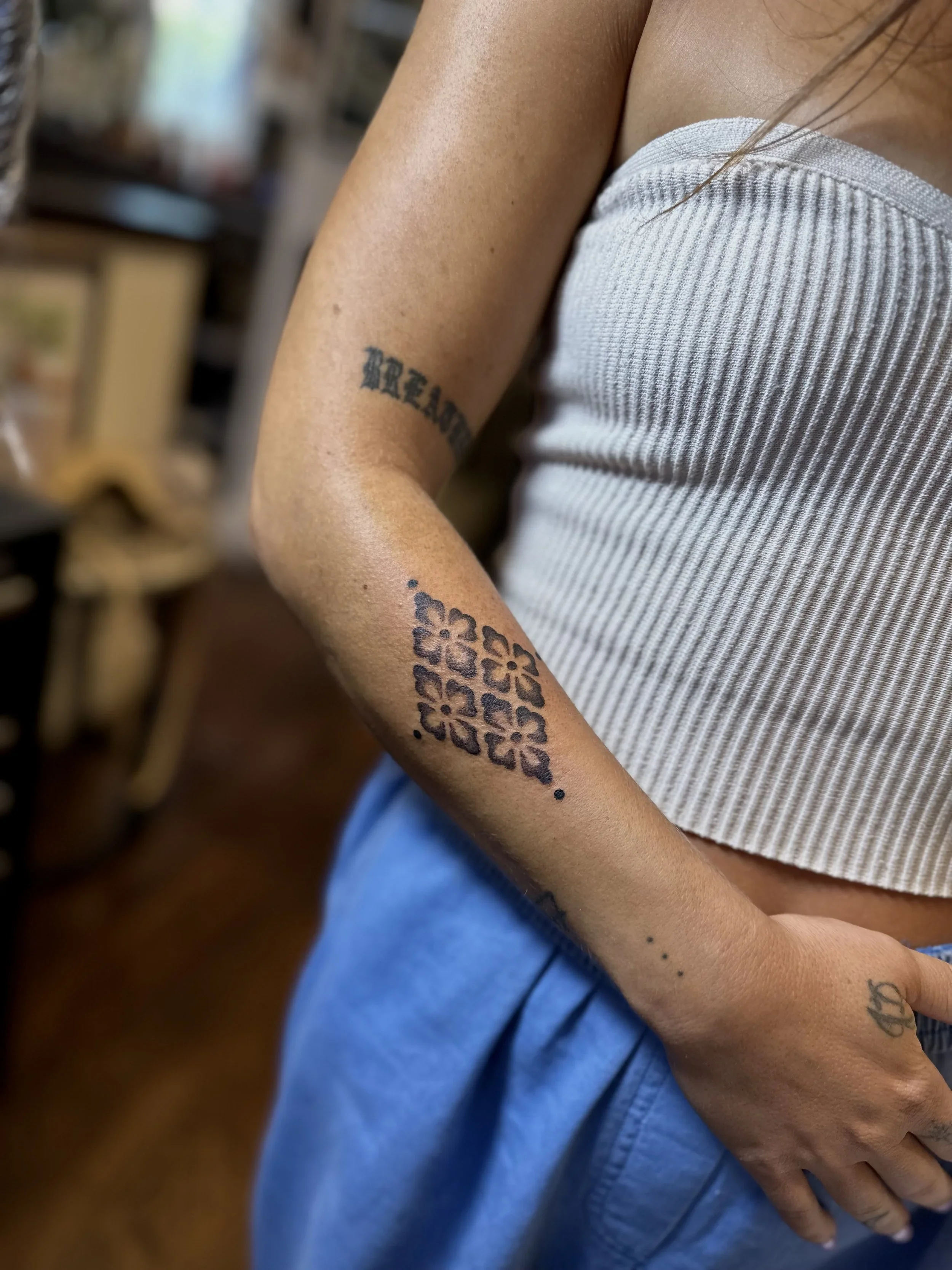 Close-up of a person's arm with a tattoo of flowers and the word 'RELACE' in black ink, dressed in a white sleeveless top and blue jeans, indoors with blurred background.