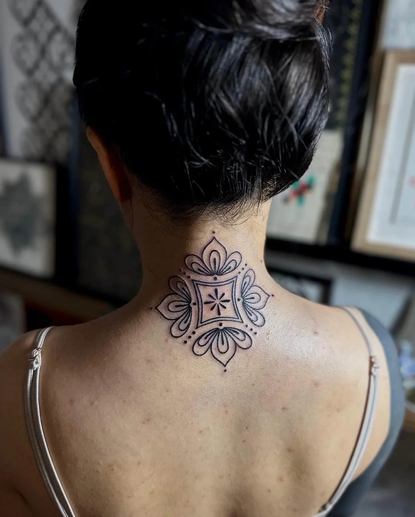 A woman with short dark hair and a detailed geometric tattoo on her neck, wearing a thin-strapped top, inside a room with framed photographs or artwork on the wall in the background.