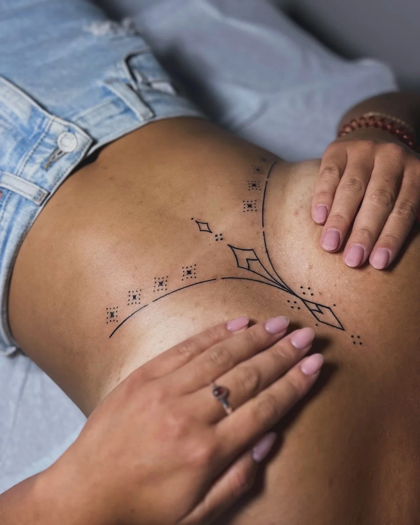 Close-up of a person's lower back with a black dotted line and geometric tattoo design, wearing denim shorts, with two hands touching the skin.