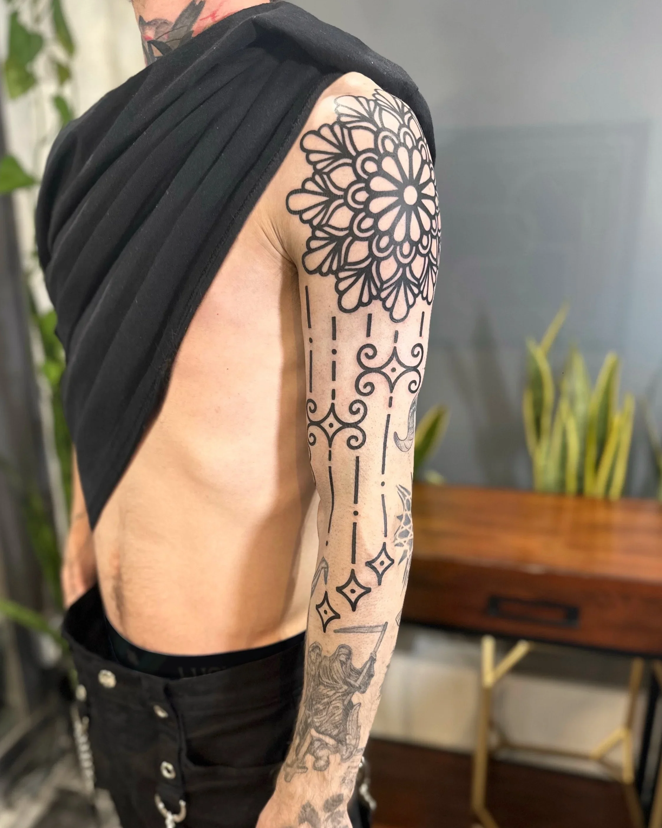 Close-up of a person's upper arm and shoulder displaying a large, black geometric and floral tattoo sleeve. The person is wearing a sleeveless black shirt, and there are additional tattoos visible on the neck and chest area. The background is indoor 