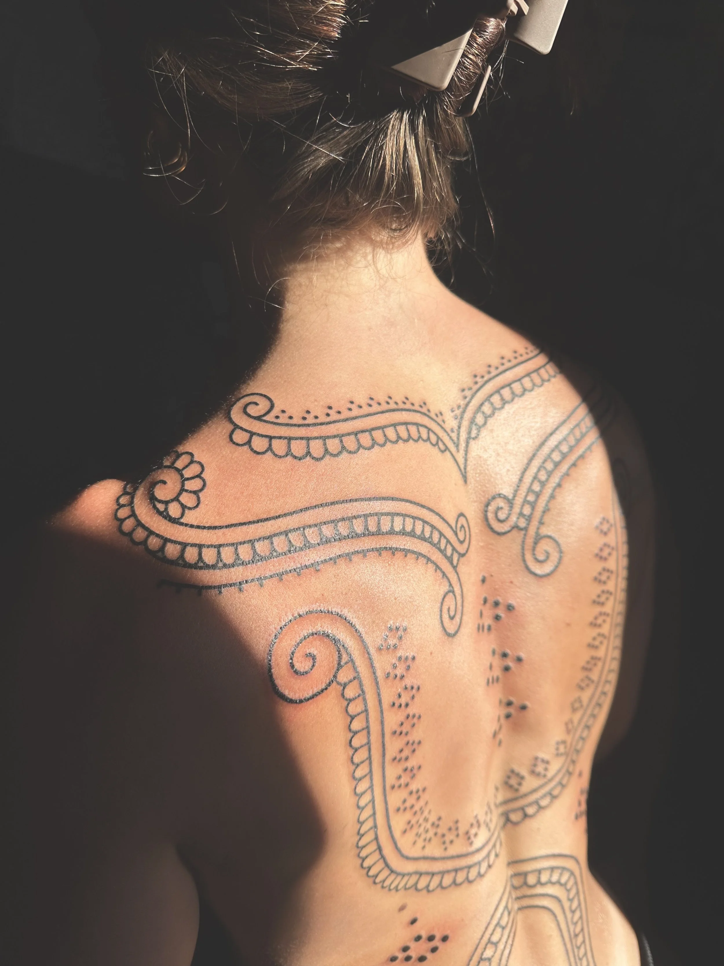 Close-up of a woman's shoulder and back showing an intricate black tattoo design with swirls, dots, and patterns, and part of her brown hair secured with a brown hair clip.