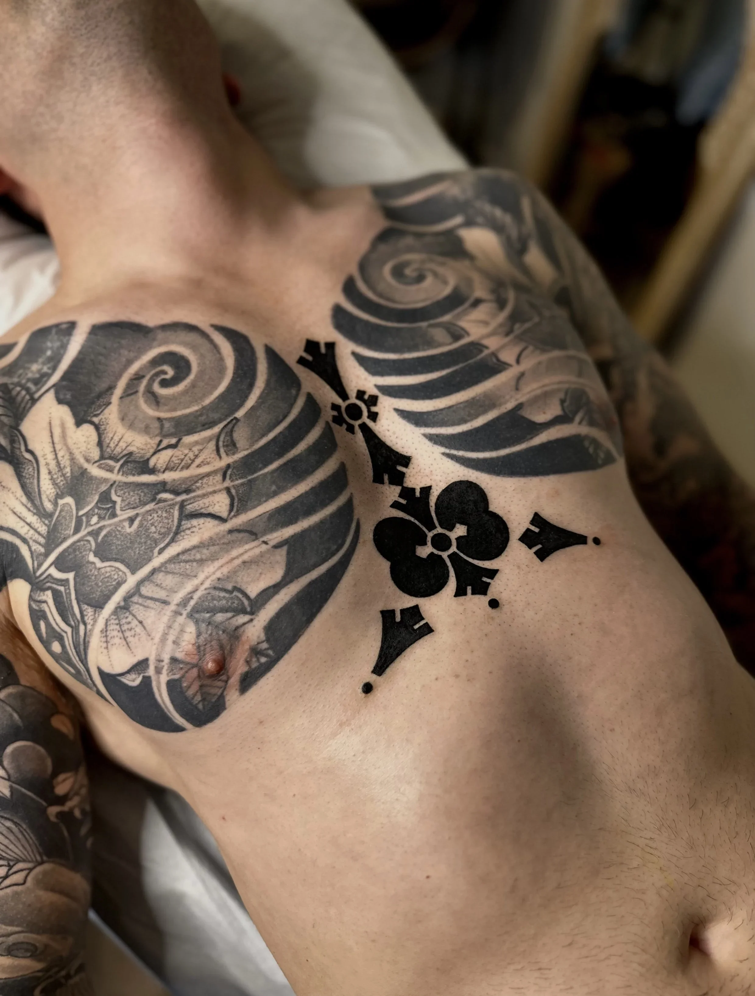 Close-up of a person's upper body with tattoos, including a large sleeve tattoo with swirling patterns and flowers, and a black design on the chest and shoulder area.